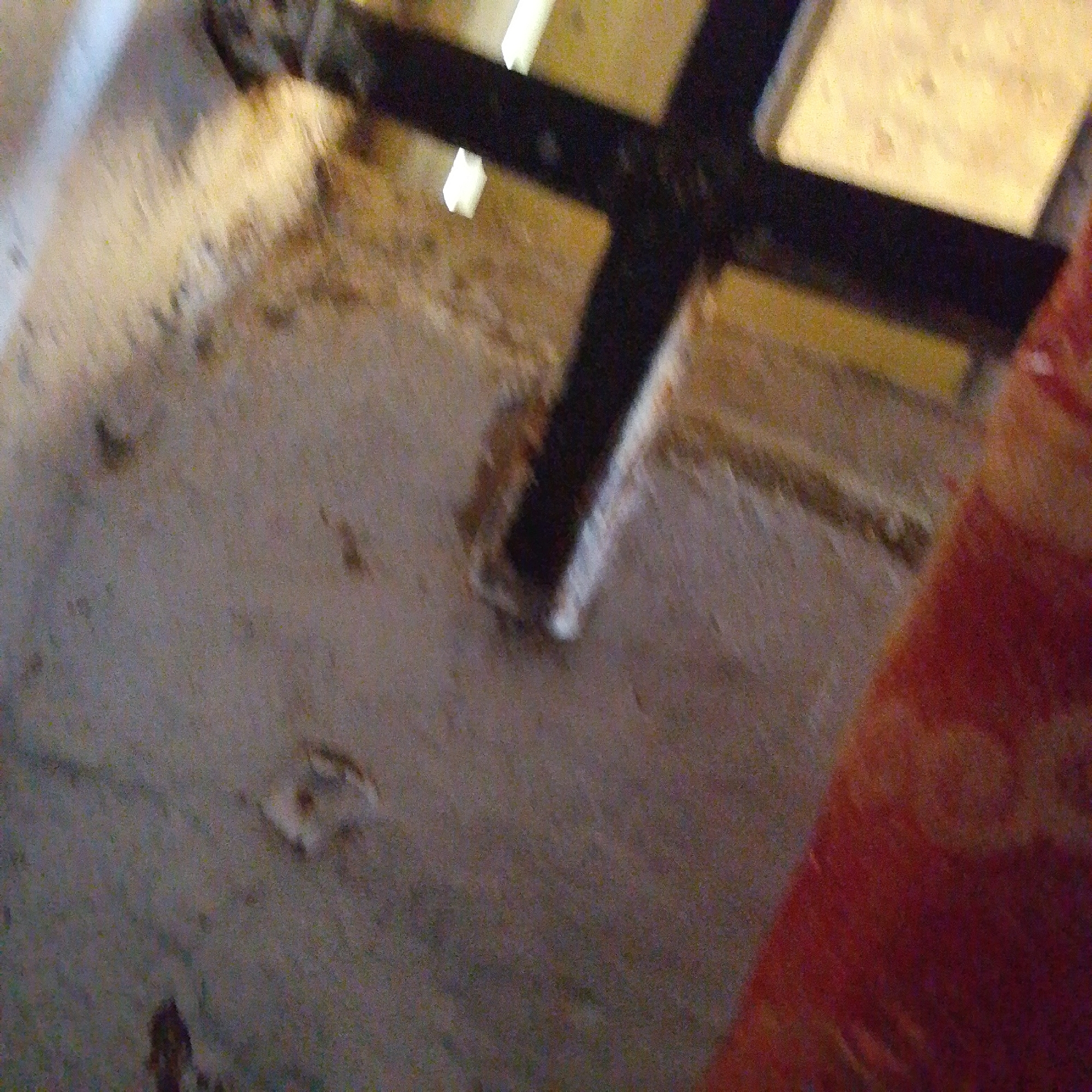 The image shows a section of what appears to be a ceiling or roof structure, possibly in an indoor or covered outdoor area. A dark, cross-shaped beam is prominent, with a lighter yellow or beige material visible above and around it. The surface below the beam is a textured, grayish-brown material, possibly concrete or a similar construction material. There are some darker smudges and lighter splotches on this surface, suggesting it is not entirely clean. On the right side of the image, a bright red and orange textured object is partially visible, which could be fabric or another material. The overall impression is of an unfinished or utilitarian structure. Without additional context or clearer details, it is not possible to determine the specific location within Jalingo, Nigeria.