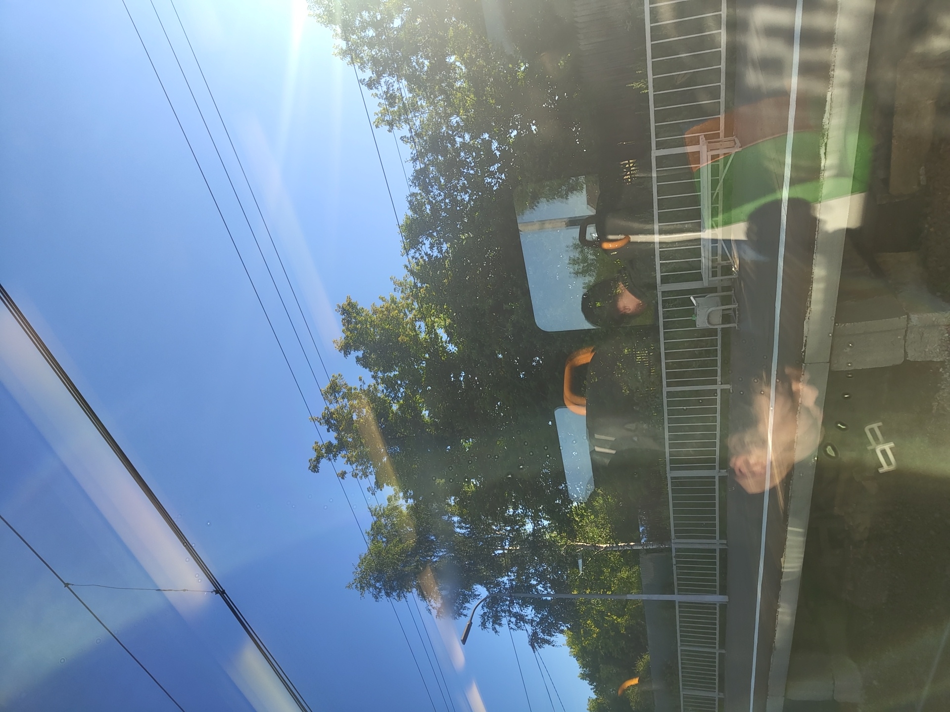 This image depicts a view from a train window, likely at a train station in Konobeevo, Russia, on a clear, sunny day. The sky is a vibrant blue, with several overhead wires visible. Lush green trees line the background, with some appearing to be birch trees due to their slender trunks and lighter foliage.
In the foreground, the train platform is visible, bordered by a white metal fence. Several benches are present on the platform, some with orange accents. Reflections on the train window obscure some details, but the silhouette of a person wearing a dark cap is visible, along with the top of a head with dark hair. Other reflections suggest the presence of more people and possibly parts of the train's interior. The platform surface appears to be made of concrete and gravel. The overall scene suggests a typical day at a suburban train station.