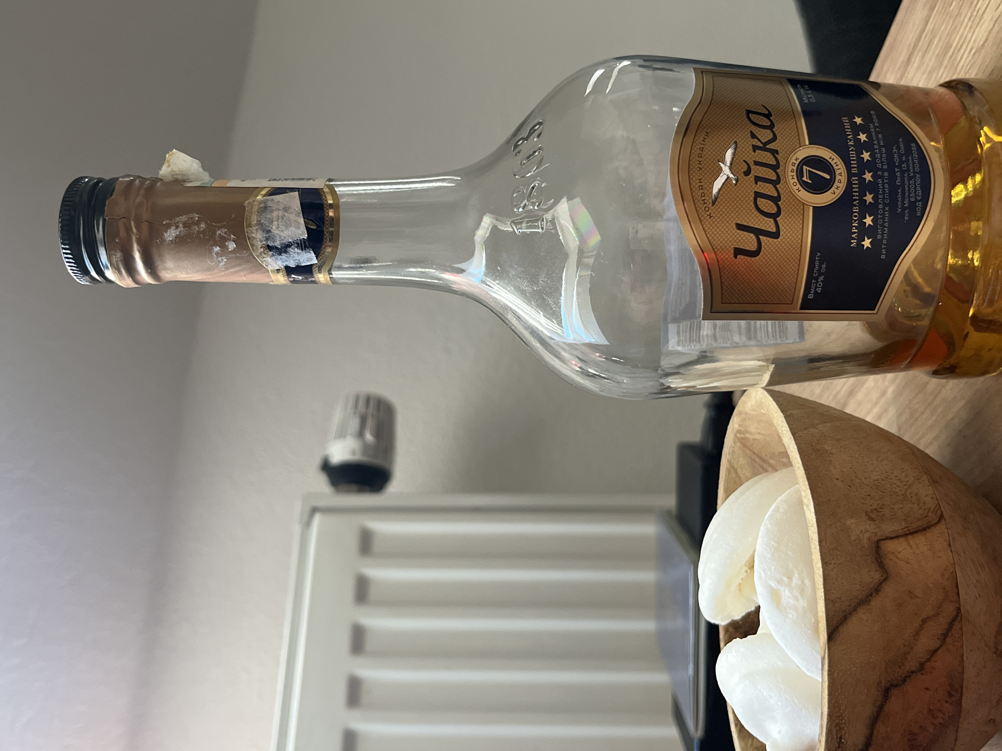 In Saint-Vith, Belgium, a close-up shot focuses on a nearly empty bottle of "Чайка" (Chaika) Ukrainian cognac, a brand known for its aging process. The bottle, with a gold and navy blue label, sits on a wooden table, reflecting light and revealing embossed numbers that appear to be "1803." Beside the bottle, a small wooden bowl contains several white, pill-shaped candies, possibly marshmallows or meringue. In the background, a white radiator is partially visible, along with a portion of a grey wall. The scene is well-lit, suggesting an indoor setting.