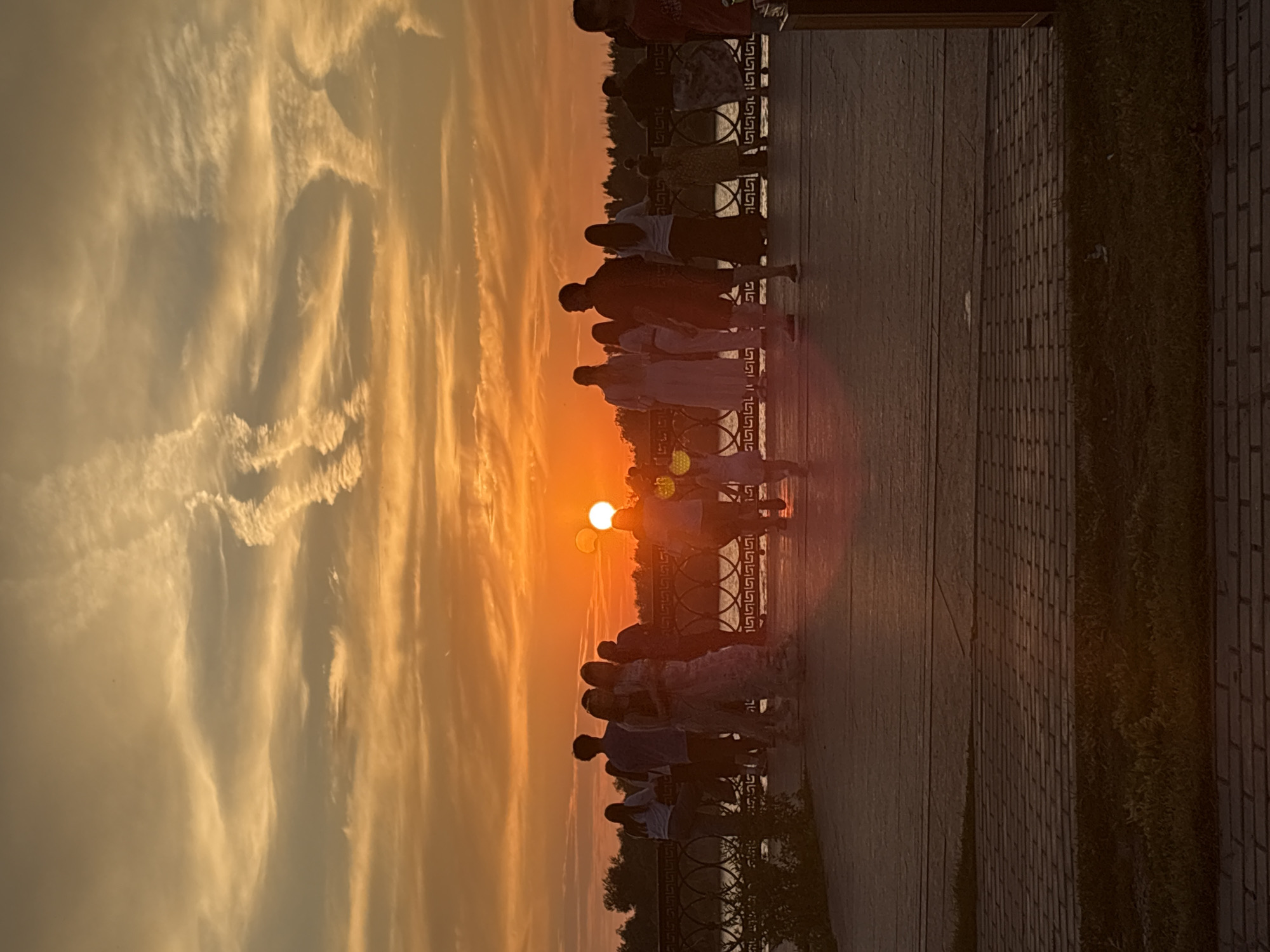 A silhouette of a crowd of people is visible on a waterfront promenade during sunset in Astrakhan, Russia. The sun, a bright orange orb, is low in the sky, casting a warm glow and dramatic cloud formations across the horizon. The people are facing towards the sun and the water, some appearing to be conversing or strolling. A decorative railing separates the promenade from the body of water, and beyond that, a treeline is visible. The foreground shows a paved walkway and some grassy areas. The overall atmosphere is tranquil and serene, characteristic of an evening by the water.