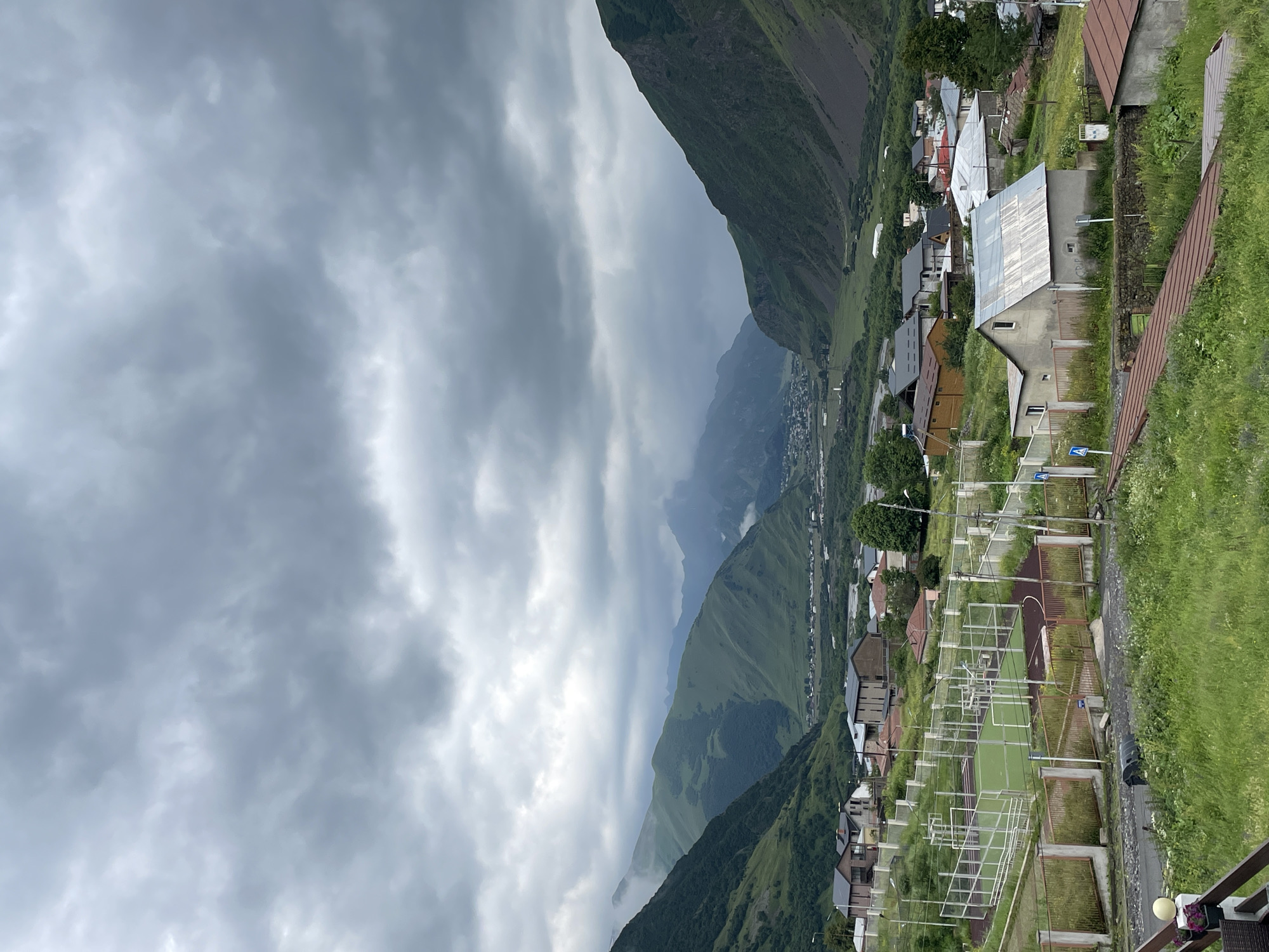 The image captures a panoramic view of a mountainous landscape, likely the town of Stepantsminda, Georgia, under a heavily overcast sky. In the foreground, lush green grass covers the terrain, with a few residential buildings nestled into the hillside. One prominent structure in the mid-ground is a single-story house with a corrugated metal roof, situated behind a low stone wall. Further to the left, a fenced sports complex, appearing to be a tennis or paddle court, is visible. The middle ground is populated with more houses, characterized by their varying architectural styles and roof materials, some with dark, sloped roofs and others with lighter, flat ones. Beyond the residential areas, the landscape dramatically rises into steep, verdant mountains that recede into the distance. The valley floor, visible between the mountains, contains a scattering of more buildings, suggesting a larger settlement. The sky is filled with dense, dark clouds, casting a somber mood over the scene and obscuring the tops of the distant peaks. There are no people or animals visibly present in the image.