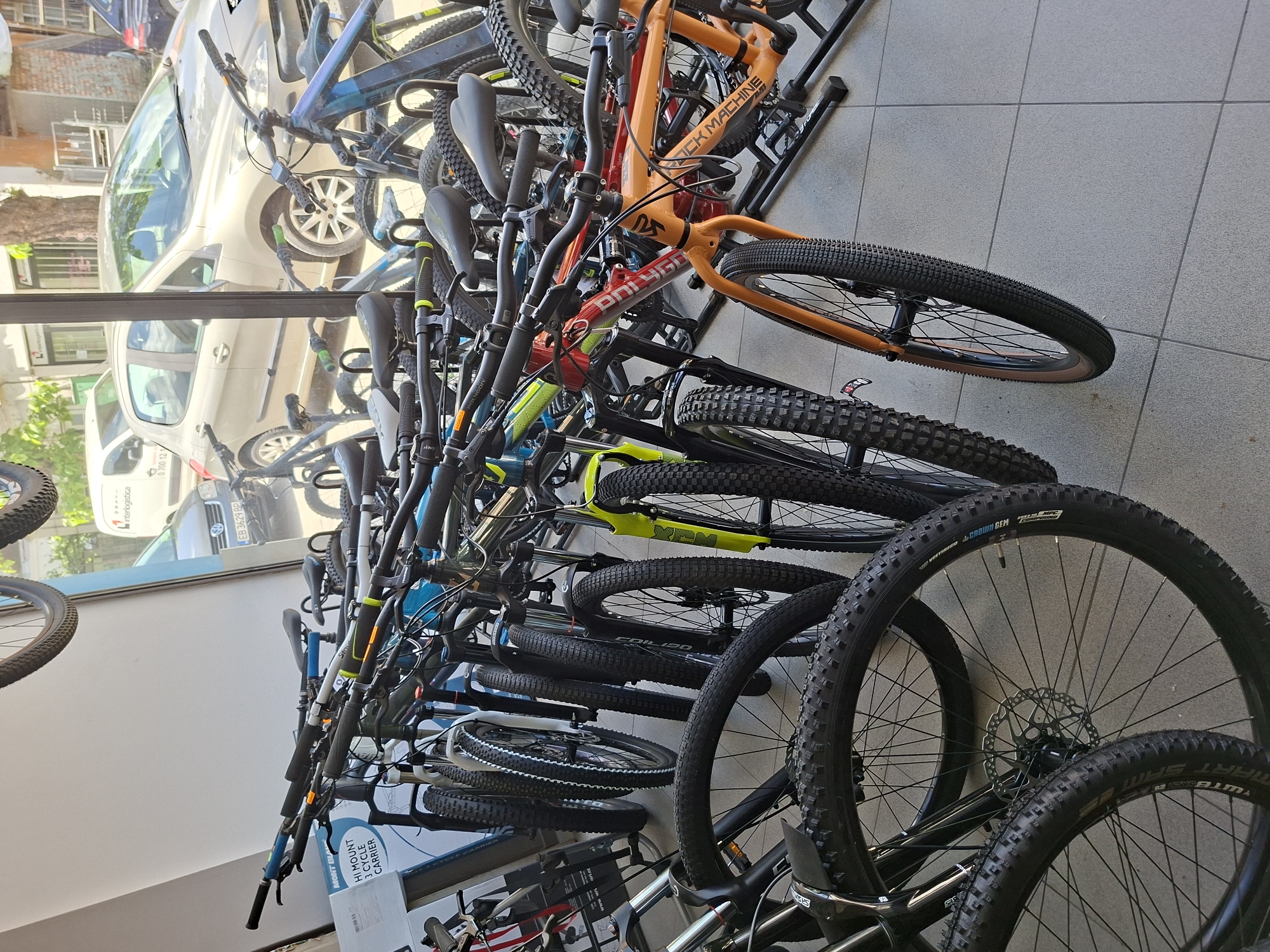 The image shows a collection of bicycles on display in a shop, likely a bicycle store. The bicycles are arranged in racks, showcasing various models and brands like "Rock Machine," "Polygon," and "YT," along with tire brands like "Vittoria Crown Gem" and "Smart Sam."
Through the glass window, a reflection of the outside is visible, revealing a street scene with cars and a building. A white van with "interlogistica" written on it, a license plate reading "EB 3629 BP," and a phone number "0700 12 1" can be discerned. Based on these details, it is reasonable to deduce that the image was taken in Gabrovo, Bulgaria, as the "EB" license plate code is associated with the Gabrovo region.
