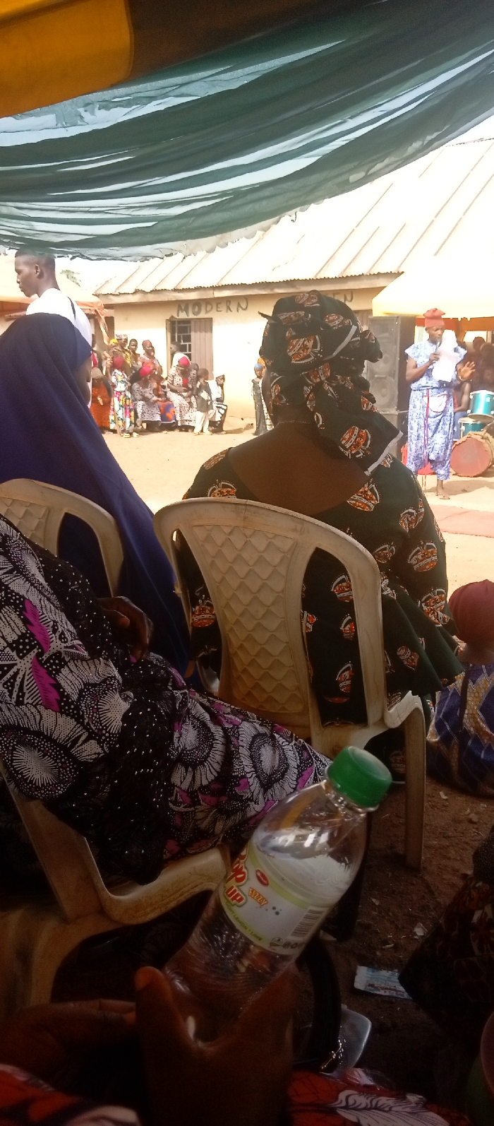 The image depicts an outdoor gathering in Kasuwan Magani, Nigeria, observed from beneath a temporary shelter constructed with green fabric. In the immediate foreground, a dark-skinned hand holds a clear plastic bottle of 7 Up soda with a green cap. The person holding the bottle is seated, partially visible, wearing a dark fabric adorned with white and magenta floral patterns. To their right, another individual is seated with their back to the viewer, wearing a dark green fabric with orange crown-like patterns and a matching headwrap. To the left, a third seated individual is partially visible from the side, wearing a dark blue head covering over a dark garment. All visible seated individuals occupy white plastic chairs.
In the midground, a group of approximately 10-15 individuals, including adults and children, are gathered in an open, unpaved dirt area. Some are seated on the ground or low structures, while others stand, dressed in various patterned traditional attires. To the far right of this group, a woman in a light blue patterned dress and a red headwrap stands near a black speaker and a large traditional drum, holding papers, indicating she may be speaking or presenting.
The background features a single-story building with a light-colored exterior and a corrugated metal roof. The word "MODERN" is distinctly painted on the building's wall. The overall scene is brightly lit by natural light.