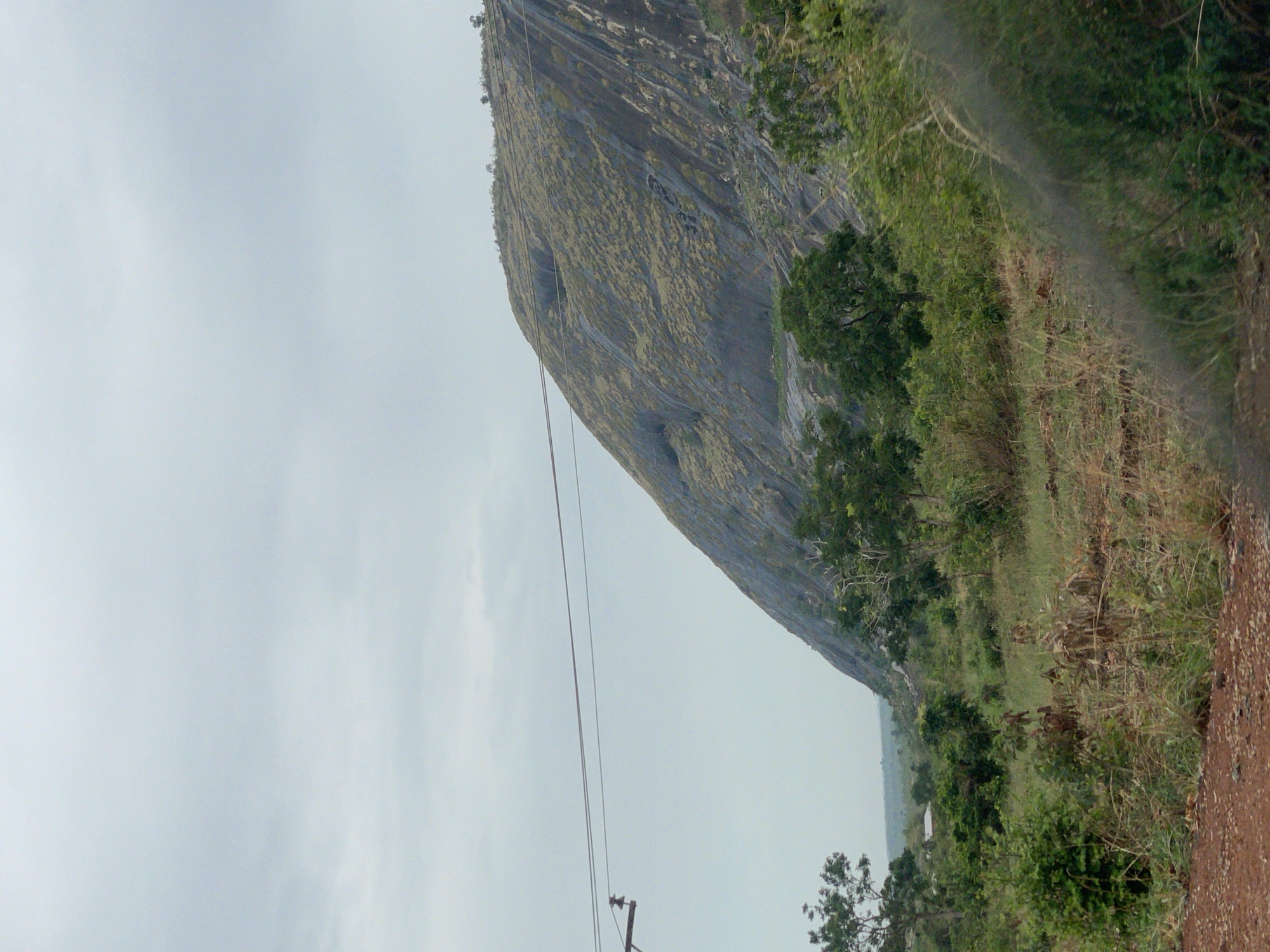 The image shows a large rock formation with a grey, weathered surface, possibly a mountain, located in Kujama, Nigeria. Patches of vegetation cover parts of the rock face. To the right of the rock formation, there is a landscape with trees and grass. On the left, a cloudy sky is visible. A utility pole and wires are positioned in the foreground. The image appears to be taken from a low angle, with the camera tilted. The immediate foreground shows a dirt or gravel surface.