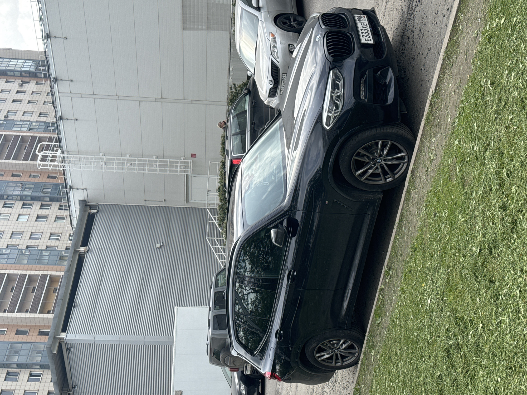 The image captures an outdoor urban scene, likely a parking area or street alongside commercial or residential buildings, under an overcast sky during daytime. The perspective is tilted, making the ground appear as a slanted surface.
The primary subject is a black BMW SUV, possibly an X3 or X4, parked near a curb with green grass. Its front license plate, visible on the grille, reads "E333XE 198". Behind it, a silver Toyota sedan (likely a Camry) is parked, displaying the front license plate "777YE 198". Further back, another light-colored car, partially obscured, shows the license plate "P7Л6P 977". All visible license plates are in the Russian format, with the "198" region code indicating Saint Petersburg, Russia.
In the mid-ground, a person is partially visible between the parked cars. The background features several multi-story buildings. One prominent building has vertical corrugated metal siding, while another, taller structure exhibits a pattern of windows and light-colored facades. A white, cage-like metal structure is attached to one of the buildings, possibly a cable management system or part of a fire escape. A red square sign with a white fire extinguisher symbol is also visible on a building. The overall scene suggests a typical urban setting with parked vehicles next to modern architecture.
