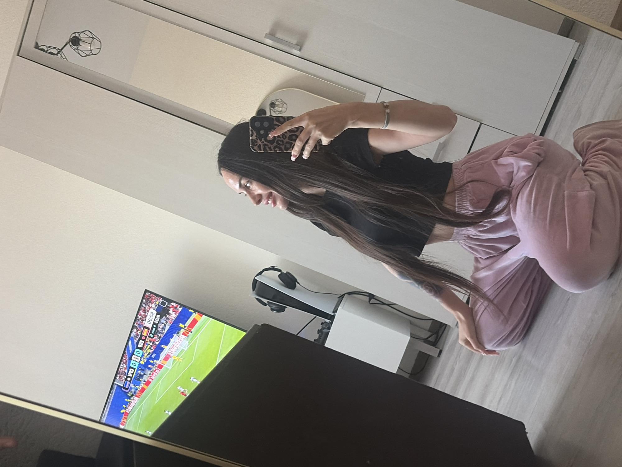 A young woman with long, dark hair is sitting on a light-colored floor, facing a television screen showing a soccer match. She is wearing a black crop top and loose pink sweatpants. The woman is holding a smartphone with a leopard-print case in her right hand, taking a selfie reflected in a large mirror to her right. The mirror also reflects a modern ceiling light fixture. To her left, a large dark furniture item, possibly a cabinet, partially obscures the television. Next to the television is a white stand holding a white gaming console with headphones placed on top, and a game controller. The room appears to be a living space or bedroom with light-colored walls and a wooden floor. The scene is indoors in an unknown urban location.