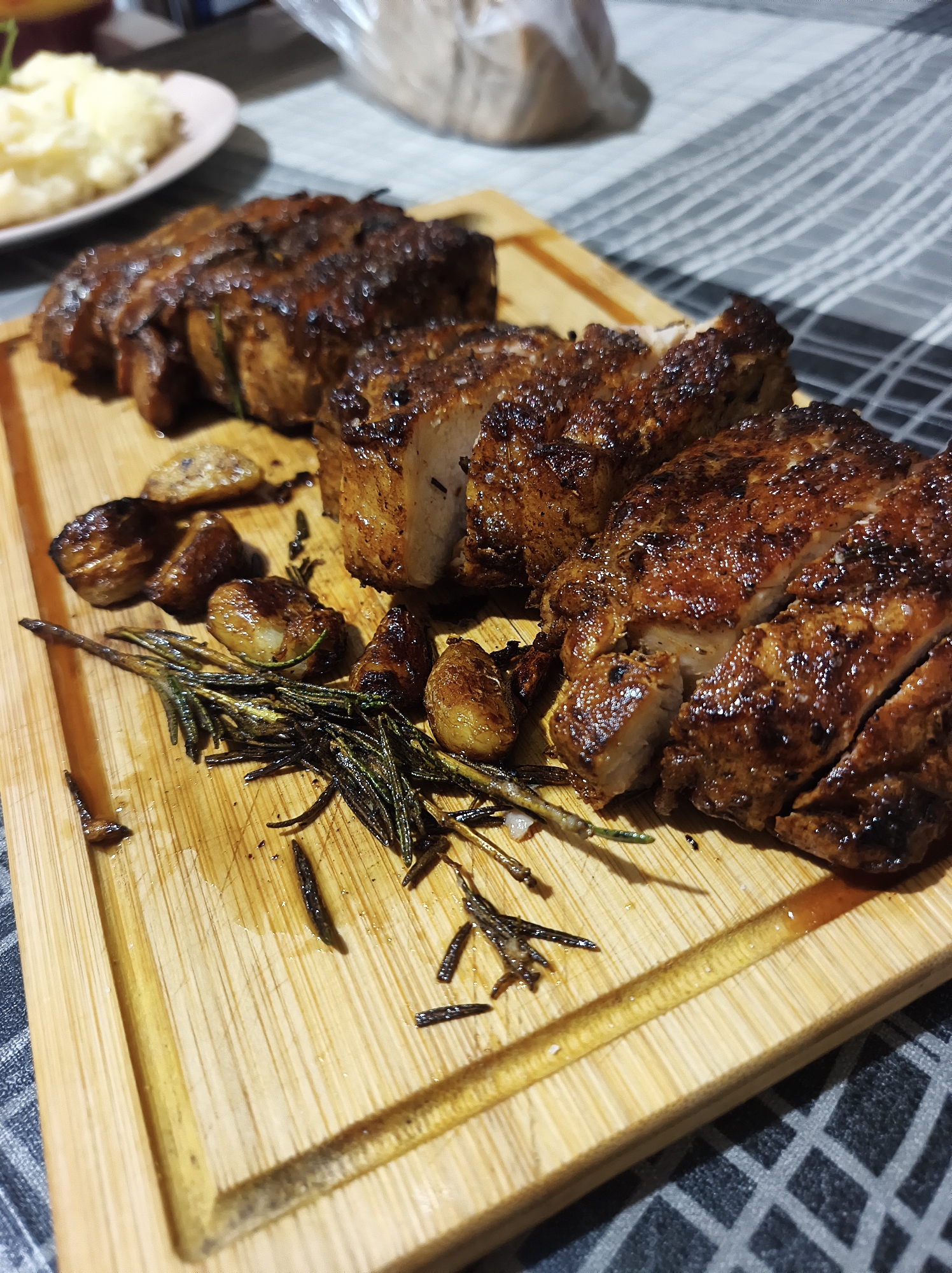 The image shows a wooden cutting board with three pieces of sliced meat arranged on it. The meat appears to be cooked, possibly roasted or grilled, and has a dark brown, slightly caramelized exterior. Scattered around the meat are roasted garlic cloves and sprigs of rosemary. In the background, out of focus, is a plate of what looks like mashed potatoes. The cutting board rests on a surface with a geometric gray and white pattern. The setting is indoors, but there's no clear indication of the specific location within Ternopil, Ukraine.