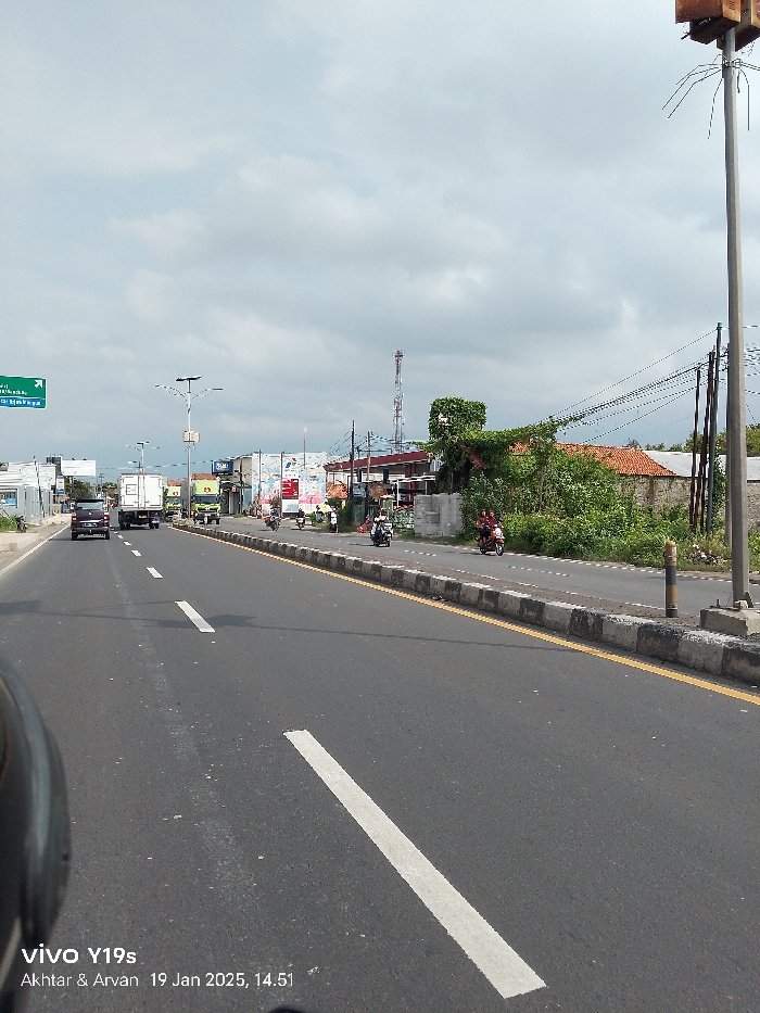 This image presents a daytime scene of a multi-lane road in Arjawinangun, Indonesia, likely captured from a moving vehicle. The road is asphalt, marked with white dashed lines separating lanes and a solid yellow line along the concrete median on the right.
Vehicles dominate the road, including several cars and trucks in the main lanes, and numerous motorcycles riding on the shoulder or side lane to the right of the median. One motorcycle prominently features two passengers. The overall activity suggests typical daily traffic.
The roadside on the right is a mix of commercial establishments with large signboards, dense green vegetation, and some residential buildings with reddish-brown tiled roofs. In the background, a tall, slender antenna tower stands out against the sky. Utility poles with numerous overhead wires line the right side of the road. A distinctive, wooden box-like structure is mounted on a pole in the immediate foreground on the right.
The sky is largely overcast with grey clouds, but patches of brighter sky indicate daylight. Shadows suggest it is mid-afternoon. A green overhead road sign on the left side of the road is partially visible, showing Indonesian place names such as "Jakarta," "Cikampek," and "Bandung."
A watermark in the bottom left corner reads "vivo Y19s," "Akhtar & Arvan," and the date and time "19 Jan 2025, 14:51."