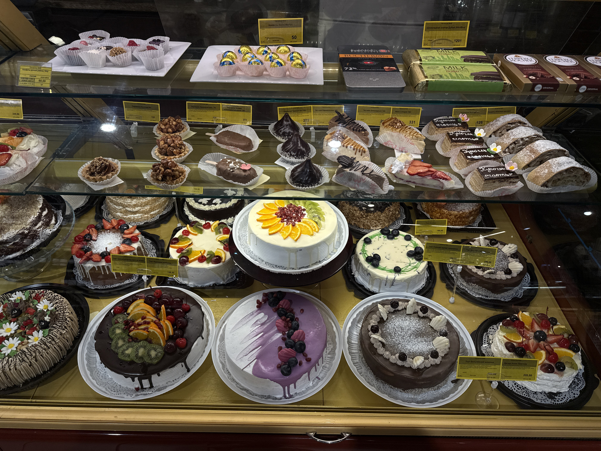 This image captures a vibrant display case filled with a wide variety of desserts and confections, likely in a bakery, cafe, or supermarket. The scene is brightly lit, showcasing an array of cakes, pastries, and chocolates arranged neatly on multiple glass shelves. No people are visible.
The top shelf features small individual desserts in white paper cups, gold-foil wrapped "Конфеты Sorini Marilyn" (Sorini Marilyn candies) priced at "50", and various boxed chocolates including "BUCHERON 72%" and "Шоколадная плитка Дубай 200/Blind" (Dubai chocolate bar 200g/Blind) for "1201", alongside "ORIGINAL" Ferrero Rocher boxes.
The middle shelf presents individual pastries such as chocolate-dipped popsicles, nut tarts, chocolate ganache figurines, fruit and cream slices, and layered cakes labeled "Кусочек счастья" (a piece of happiness), priced at "126". Other prices like "273", "331", and "136.50" are also visible.
The bottom shelf is dedicated to larger, elaborate cakes. These round cakes are decorated with fresh fruits (kiwi, oranges, berries, cherries), chocolate drizzles, whipped cream, and piped designs, some featuring edible daisy flowers. Labels identify items such as "Торт Бельгийский шоколад" (Belgian Chocolate Cake) priced at "100,00" and "Десерт Анна Павлова" (Anna Pavlova Dessert) at "220,50". Other prices, like "189,00", are also seen. Several labels bear the country of origin "РОССИЯ" (Russia) and the "EAC" conformity mark. The prices are in a currency format suggesting Russian Rubles.