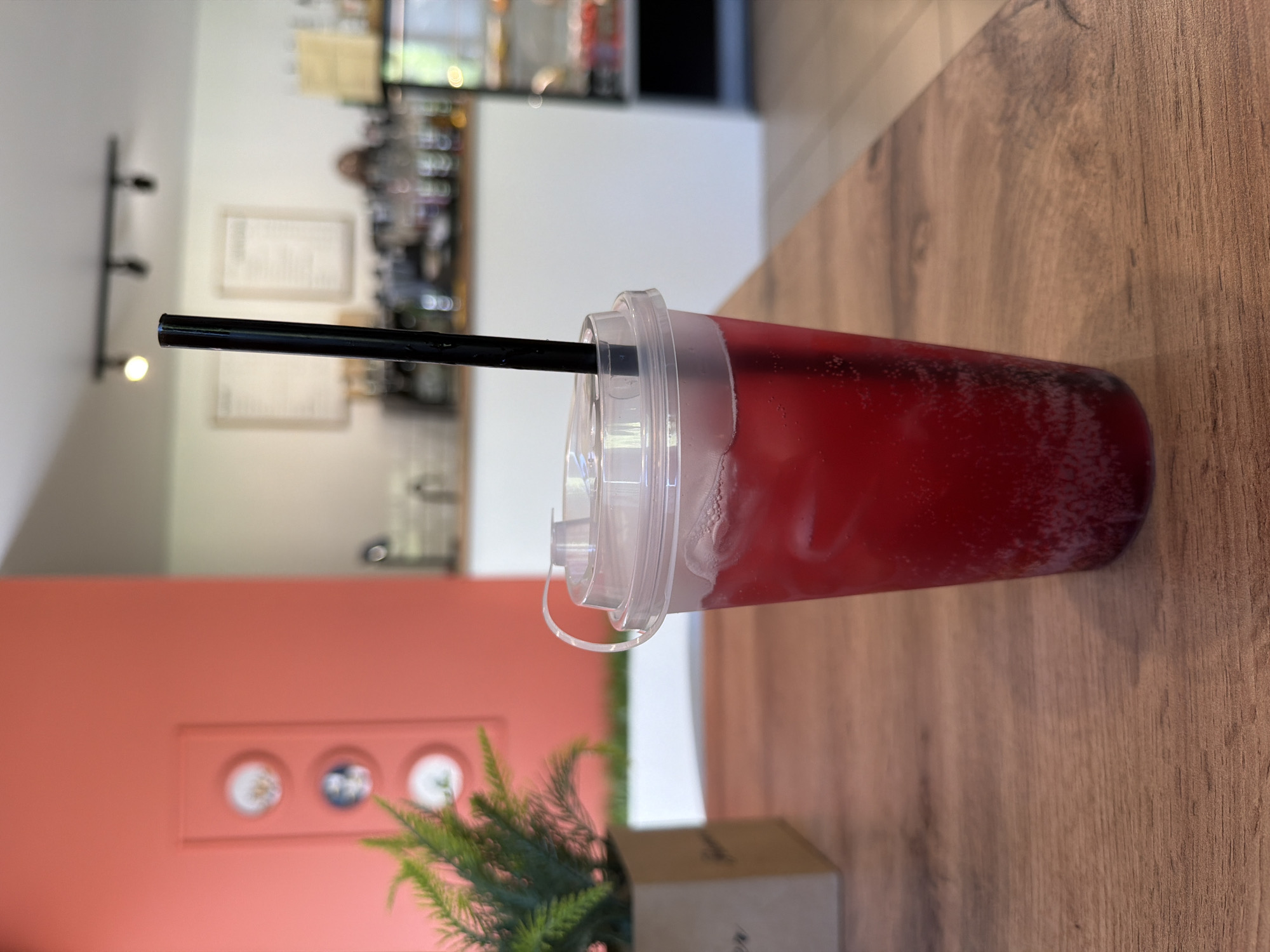A close-up, medium-shot photograph features a red beverage in a clear plastic cup with a clear plastic lid and a black straw. The cup sits on a brown wooden table, angled towards the right side of the frame. Behind the drink, a small artificial green plant in a brown pot is visible. Further in the background, a blurred interior of what appears to be a café or restaurant is visible. An orange wall with decorative white plates is on the left side. The background includes elements of a counter, shelves with items, and blurred figures of people. The lighting is natural and even. The image appears to have been taken at a café or restaurant location. The geographical location of Chaikovsky, Russia is not directly determinable from the given visual information.