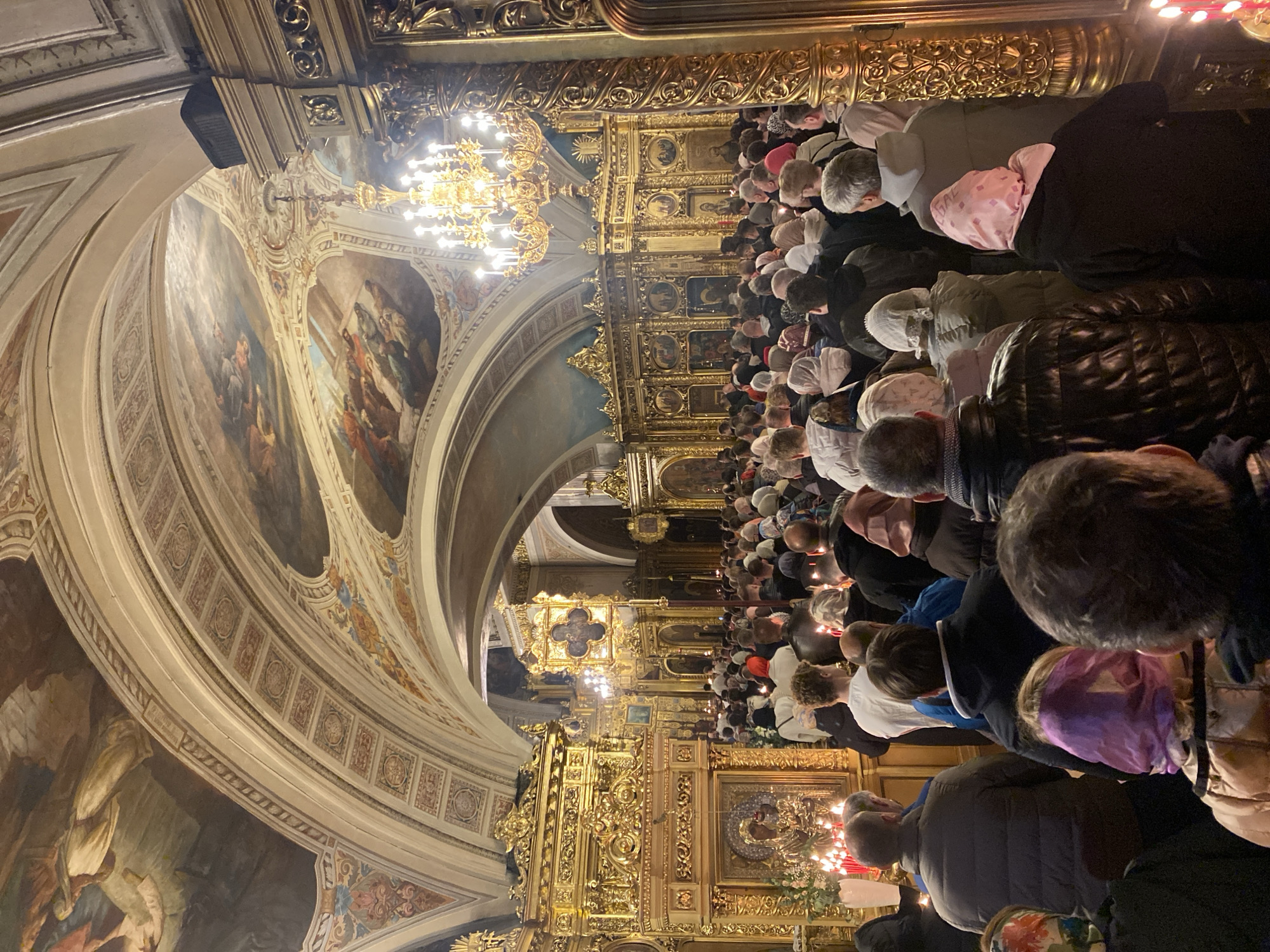 A low-angle, canted medium shot shows a packed cathedral with people facing the front with their heads bowed. The interior of the cathedral is ornate with gold leaf decor, frescoes, and lit chandeliers. The worshippers are holding lit candles. Based on visual cues such as the style of architecture, art, and religious iconography, the location is likely in the city of Moscow, Russia.