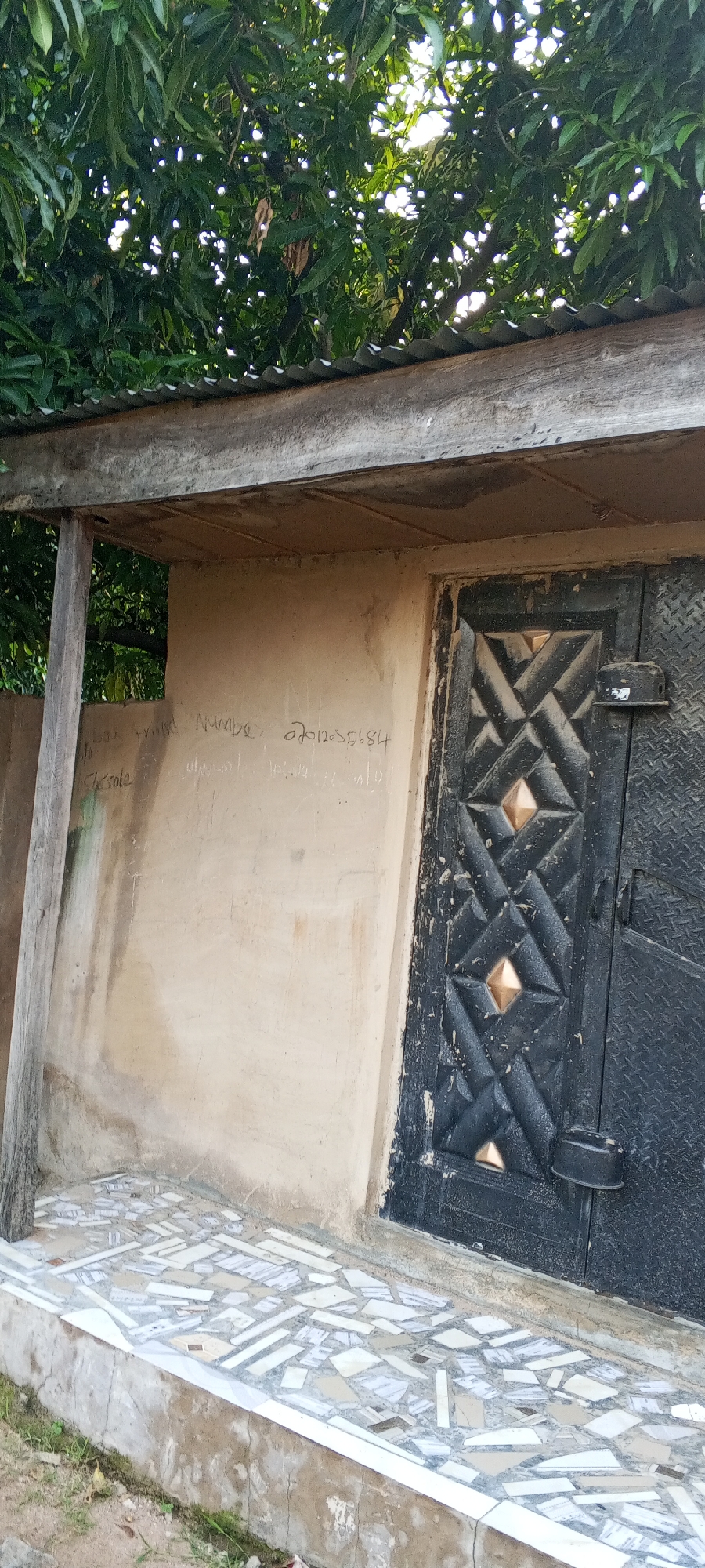 This image depicts the exterior of a building, likely a shop or dwelling, in Jalingo, Nigeria. A portion of a corrugated metal roof is visible at the top, supported by wooden beams. Below the roof, a light-colored, plastered wall is marked with graffiti. Most notably, the wall features what appears to be a name or inscription, followed by a sequence of numbers that could be a phone number: "Nuwobe 27012056184". To the right of the wall is a dark metal door with a geometric pattern, featuring diamond-shaped metallic embellishments. A latch or lock mechanism is attached to the door. The ground in front of the building is covered with a mosaic of broken tiles, laid on a concrete base. Green foliage from a tree is visible overhead. The overall scene suggests a modest urban setting.