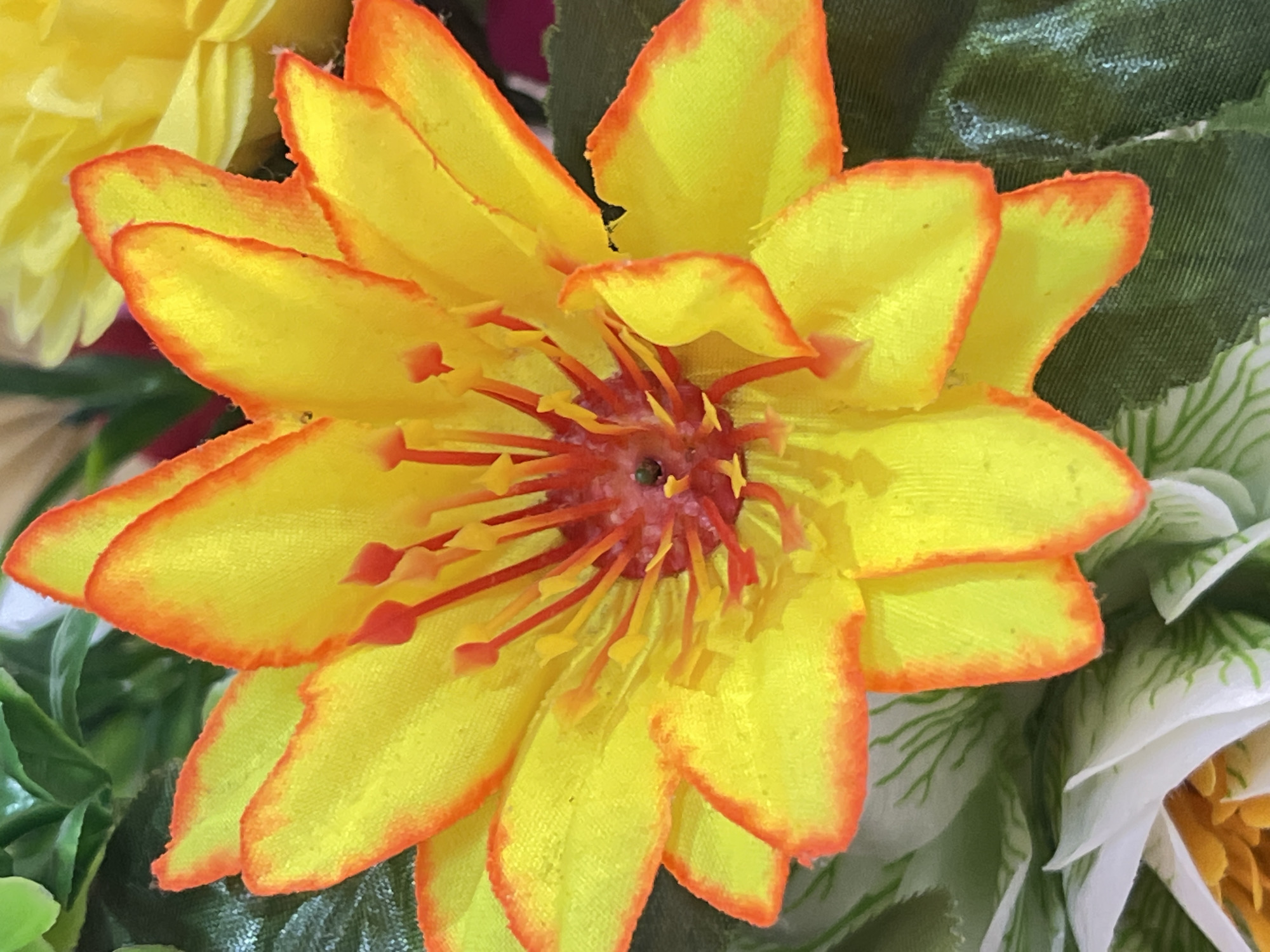 The image is a close-up, eye-level shot of a bright yellow artificial flower with orange-red edges. The flower has multiple pointed petals arranged in layers. In the center, there are numerous red stamens with yellow tips. The flower appears to be part of an artificial floral arrangement, with glimpses of green artificial leaves and other flowers, including a white one at the bottom left and a yellow one in the upper right background. The lighting is bright and even, creating a slightly soft focus effect. The scene is indoors, possibly in a decorative setting. There is no indication of the flower's specific location in Denpasar, Indonesia.