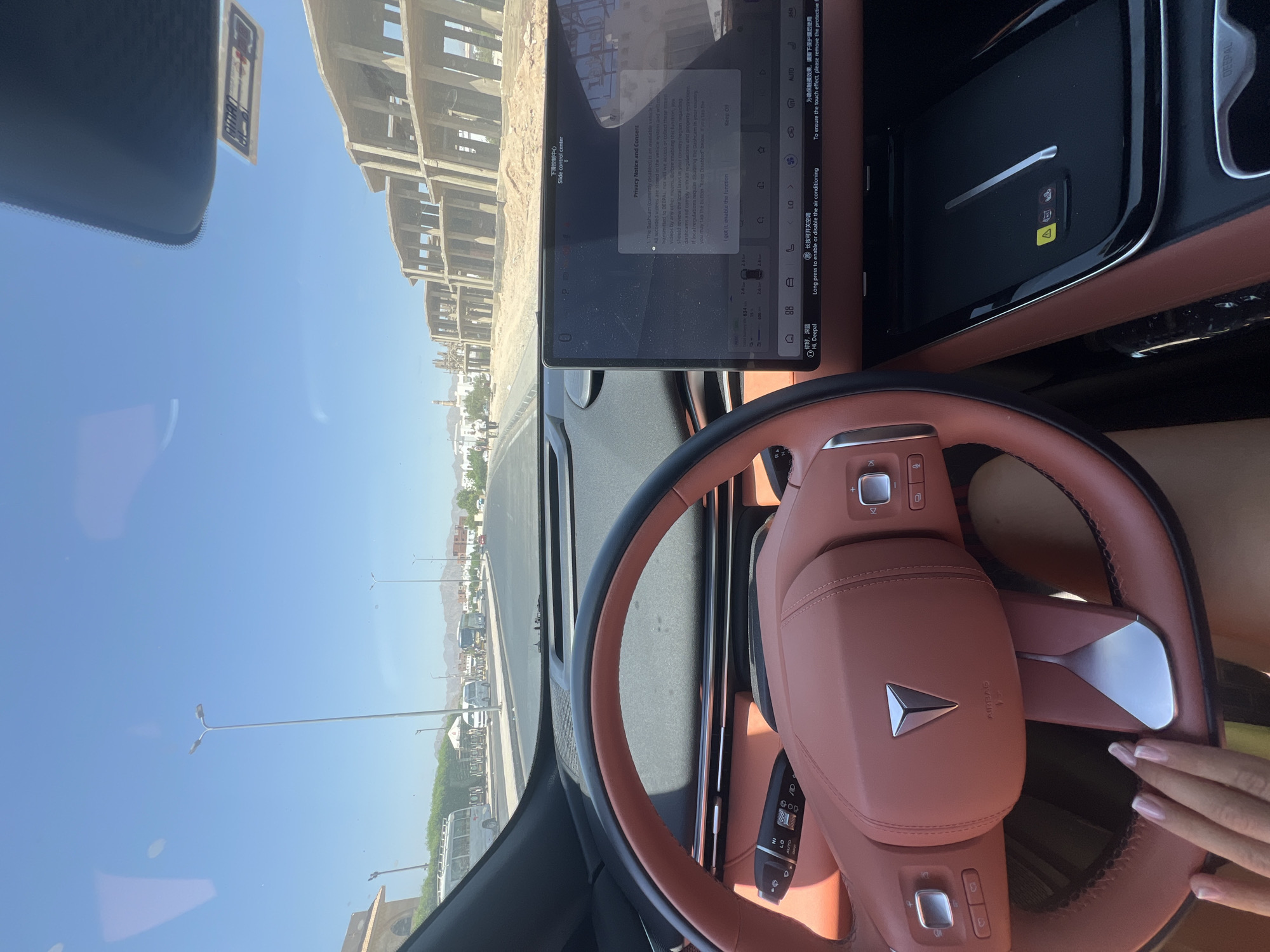 The image is a first-person perspective from inside a car in Sharm El Sheikh, Egypt. The car features a prominent tan-colored steering wheel with a metallic emblem, and a large central touchscreen display. The touchscreen shows a "Privacy Notice and Consent" pop-up in English, with options to "I got it, enable the function" and "Keep Off." Below the touchscreen, a black surface with a wireless charging pad is visible. The car is on a paved road, with a clear blue sky overhead. In the background, there are several multi-story buildings under construction, as well as smaller white buildings that appear to be residences or businesses. On the side of the road, there are streetlights, some vehicles including a white bus, and trees. The overall scene depicts a sunny day in a developing area of Sharm El Sheikh, Egypt.