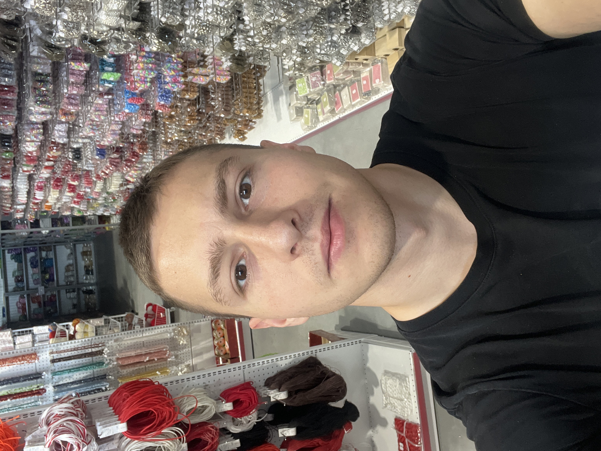 A man is taking a selfie in what appears to be a craft or bead store. He is wearing a black t-shirt and is positioned in front of shelves stocked with beads, threads, and other craft supplies. The background shows a variety of colorful items commonly found in craft stores. The image does not contain enough information to determine if the location is Burgas, Bulgaria.