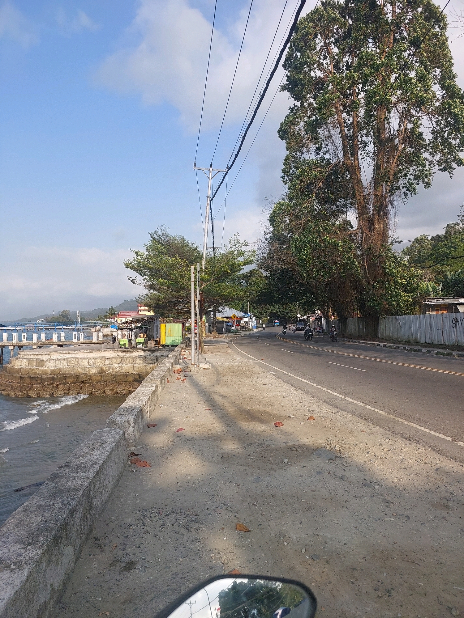 The image depicts a roadside scene in Ambon, Indonesia, likely near the coast. On the left side, a concrete seawall runs alongside the water, with waves gently lapping against it. Beyond the seawall, a pier extends into the sea, with several small buildings and structures visible on it. A road curves through the scene, with several motorcycles being ridden on it. Large trees with dense foliage line the right side of the road, with some buildings and a corrugated metal fence visible in the background. Overhead, power lines crisscross the sky. The overall impression is of a coastal road on a sunny day, with the presence of both natural and man-made elements typical of a developed area in Indonesia.