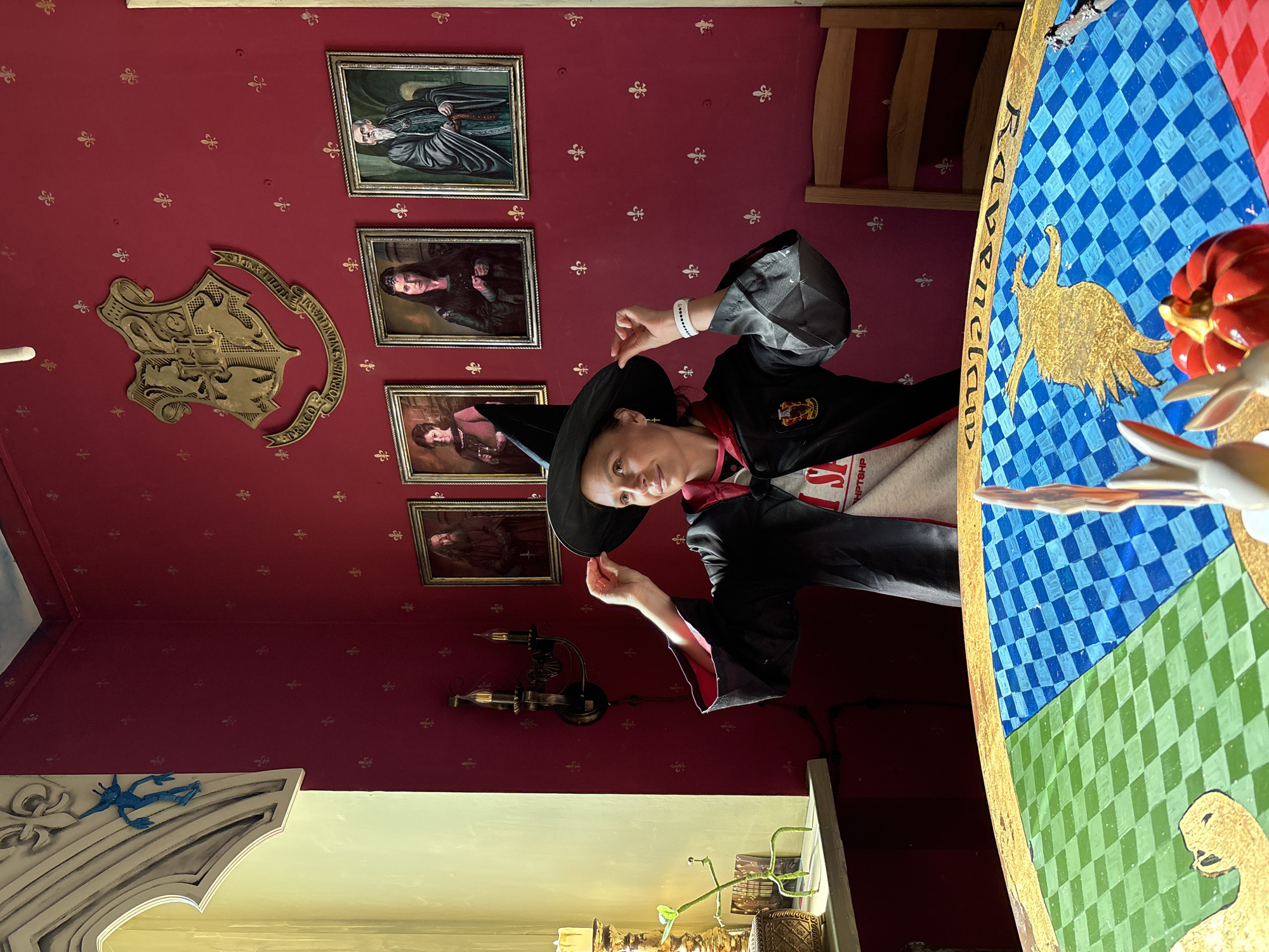 A person dressed as a witch is posing in a room with Harry Potter-themed decor. The room features red walls adorned with gold fleur-de-lis patterns, as well as framed portraits of figures from the Harry Potter series, and a decorative Hogwarts crest. A table with a checkered pattern in blue and green is in the foreground, with a golden raven and a badger on the surface. The person is wearing a black witch's hat and a black robe. A wall fixture with candles is present in the room. A portion of a shelf or cabinet with a white border is visible in the lower left, decorated with a blue figure. The location is likely a themed room or establishment that is inspired by the Harry Potter universe. The user's request includes location of Kazan, Russia. The image does not contain the information if the location of the photo is Kazan, Russia.