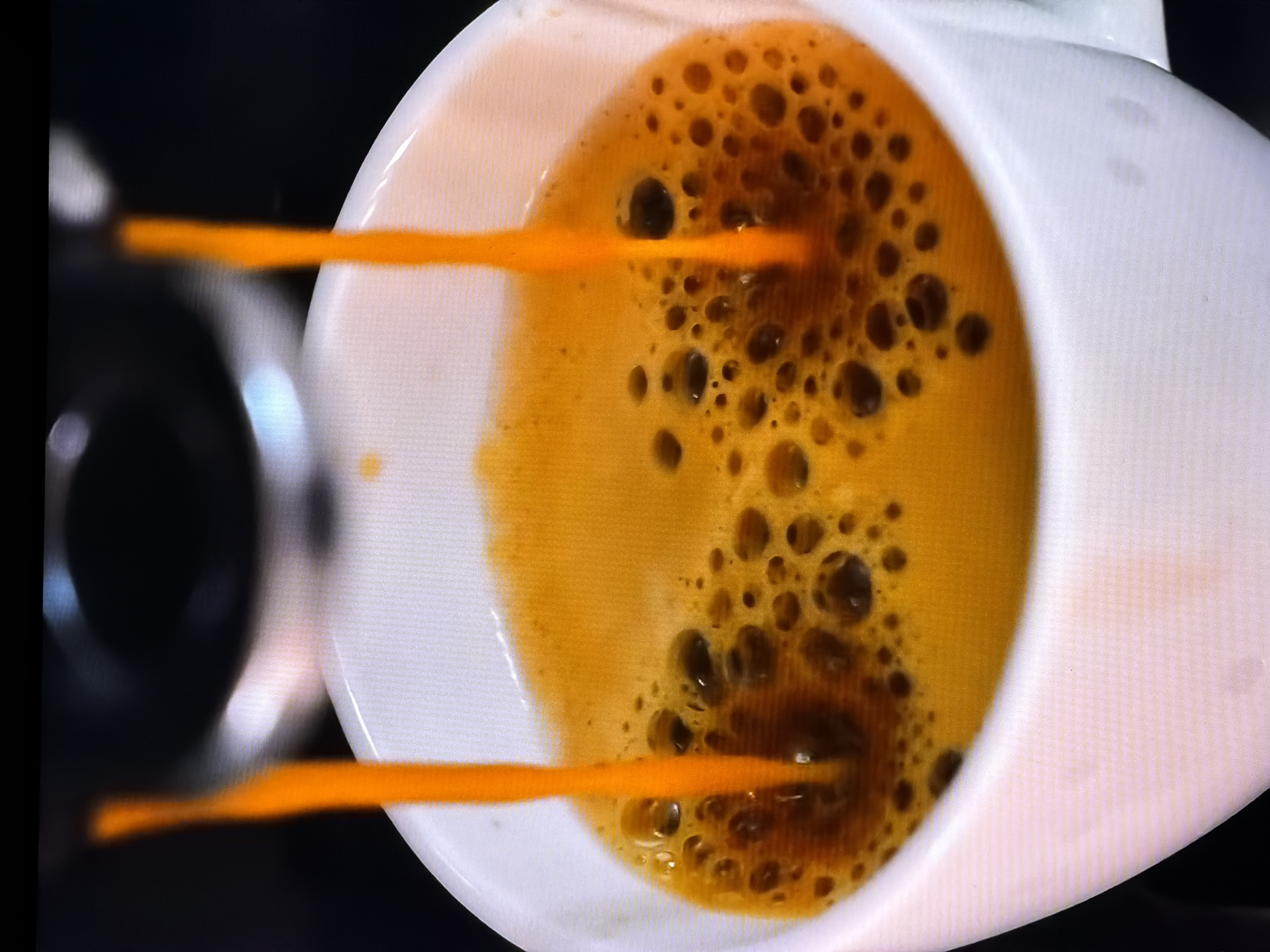 A close-up, eye-level shot shows fresh espresso being poured into a white ceramic mug. The espresso is a rich, golden-orange color and is being dispensed from an unseen machine above. A thin stream of the dark liquid cascades into the mug, creating a frothy layer of crema on top, characterized by small, dark bubbles. The mug is partially filled, with the liquid level rising towards the rim. The background is dark and out of focus, with hints of metallic or dark plastic suggesting the espresso machine. The image is captured from a perspective that emphasizes the pouring action and the visual appeal of the freshly brewed coffee. The location in Chirchik, Uzbekistan, is not depicted or suggested by any visual elements in the image.