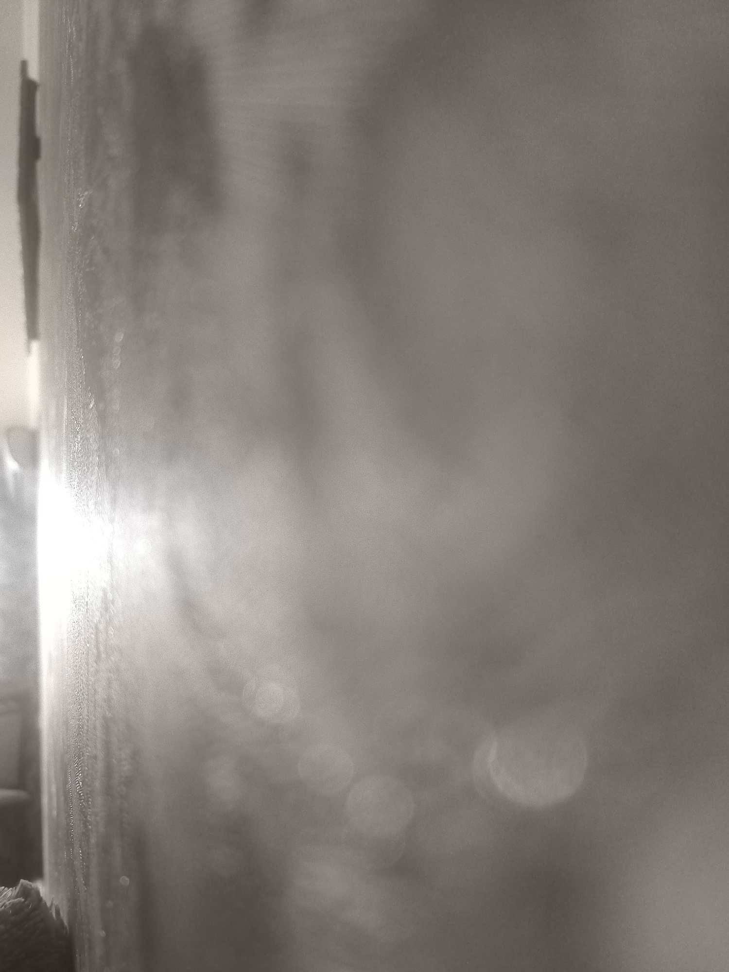 The image is a close-up, low-angle shot of a textured, possibly concrete or plaster wall. A strong, bright light source is visible on the left side of the frame, casting reflections and highlighting the rough texture of the wall. In the foreground on the bottom left, a dark, woven object, possibly fabric or netting, is visible. The background is out of focus, showing lighter tones and some bokeh effects that suggest distant lights or reflections. The overall impression is abstract and textural, with a focus on light and shadow playing across the surface. The image is in grayscale. It is not possible to determine the location in Kokand City, Uzbekistan from this image.