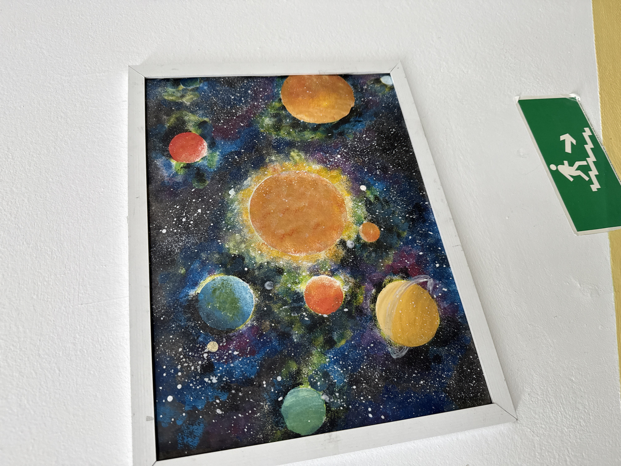 The image shows a framed painting of a solar system on a white wall. To the right of the painting is a green sign with a white symbol of a person walking down stairs. The painting is composed of several planets and stars painted with various colors. The location is in the city of Chaikovsky, Russia.