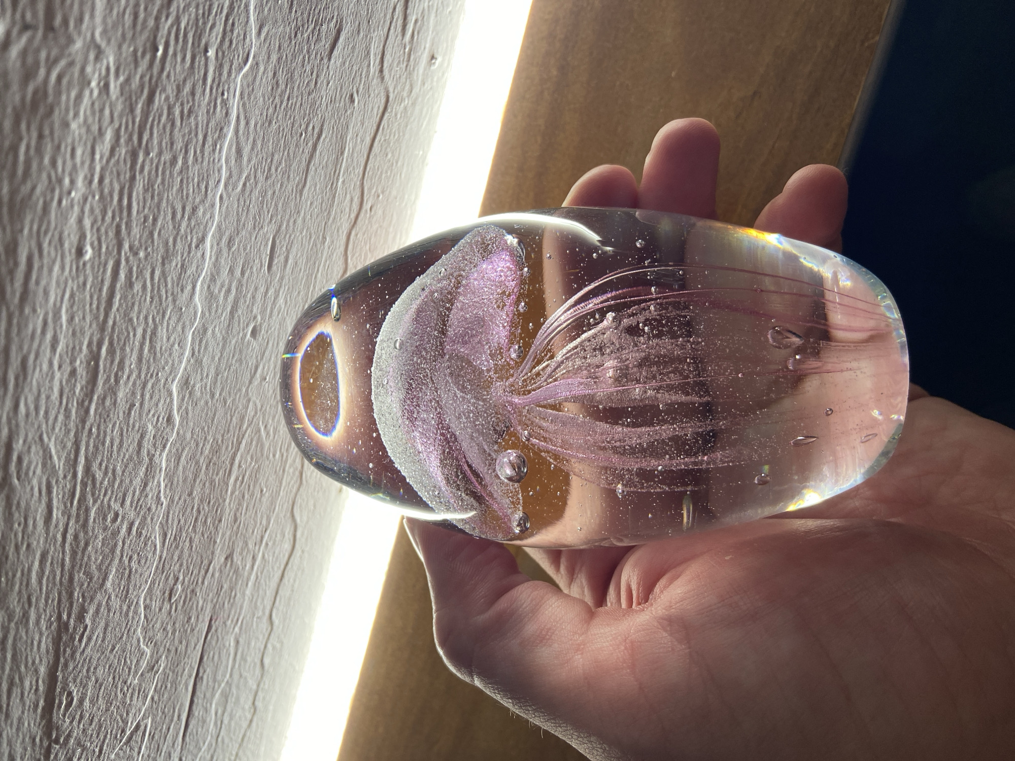 A hand holds an oval-shaped glass paperweight. Inside the glass, there is a stylized, pink jellyfish-like design. The background includes a textured white wall and a light fixture. Part of a wooden surface is visible near the hand. The image was not taken in Petrovac centar, Montenegro.