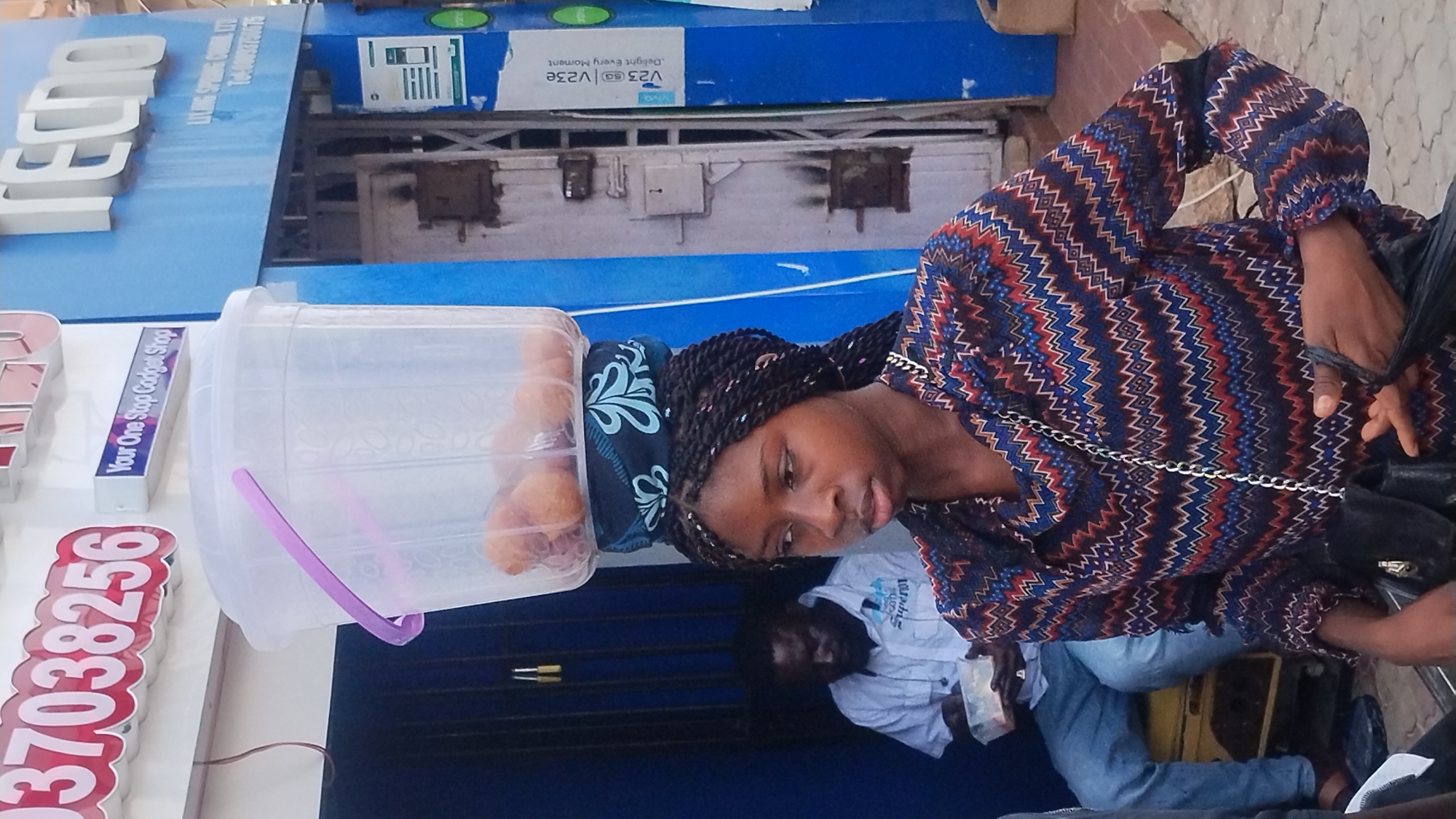 A woman stands with a clear plastic container on her head, likely filled with food items for sale. The container has a purple handle. She is wearing a patterned long-sleeved shirt and has braided hair. The location appears to be a business district, indicated by signs in the background. Prominent is a "Tecno" sign and another sign that reads "Your One Stop Gadget Shop" with the number "037038256". A man sits behind her, and parts of other people are visible. Based on the language on the signs, context clues about the surroundings, and common vendors, the location is likely in Jos, Nigeria.