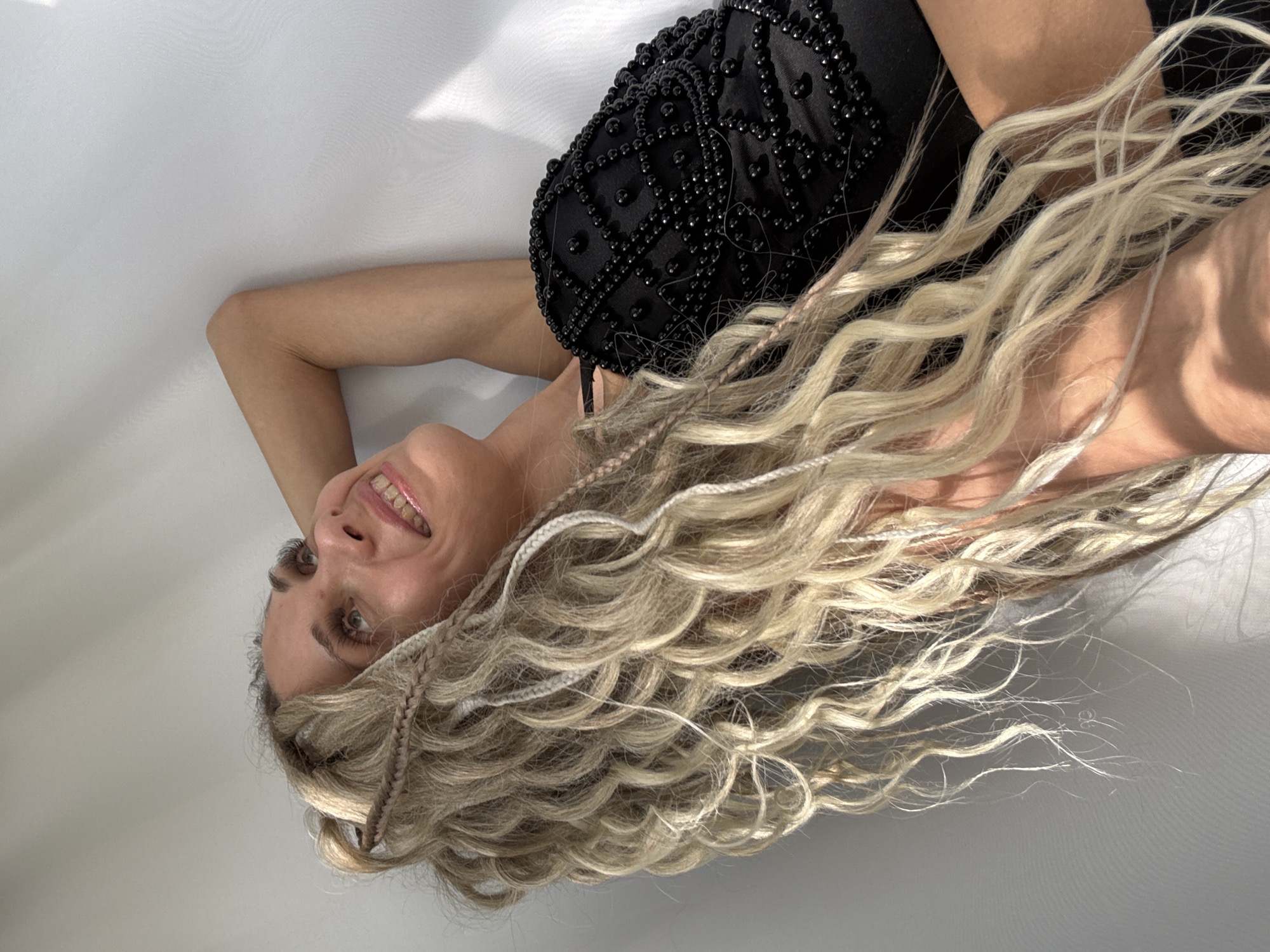 A woman with long, wavy blonde hair with braided sections lies on a white surface, seemingly a studio backdrop. She is wearing a black dress with beaded detailing. Her head is slightly tilted, and she is looking upwards with a smile. Her skin tone appears fair. The lighting is bright and casts soft shadows on the white surface. There is no indication of the location being in Rusanovo, Russia.