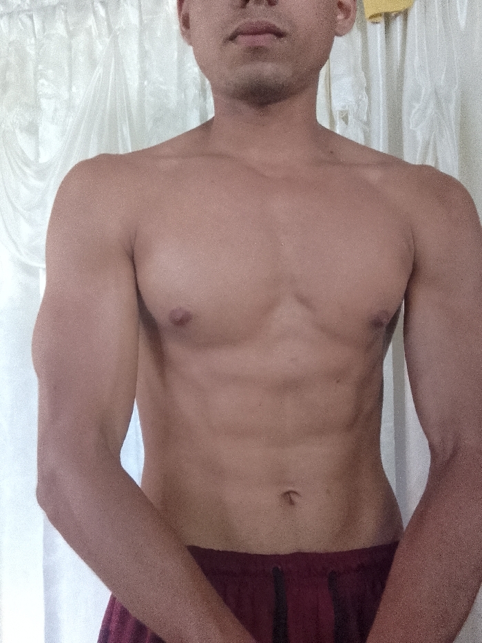 The image displays a male individual, partially cropped from the mid-chest to the upper thighs, standing shirtless. The subject exhibits a light brown skin tone and a muscular physique, with visible definition in the pectoral muscles and a discernible outline of the abdominal muscles. His face is partially visible, showing from the nose downward. He is dressed in dark red athletic shorts featuring a dark-colored drawstring or stripe at the waistline. Both arms are bent at the elbows, positioned with the forearms angled slightly inward towards the center of his body. The background consists of a light-colored, possibly white or off-white, translucent curtain with prominent vertical folds. A small, indistinct yellow object is partially visible in the upper right quadrant of the background. The lighting is even, consistent with an indoor environment. There are no discernible visual cues within the image to specify the location as Juan Griego, Venezuela.