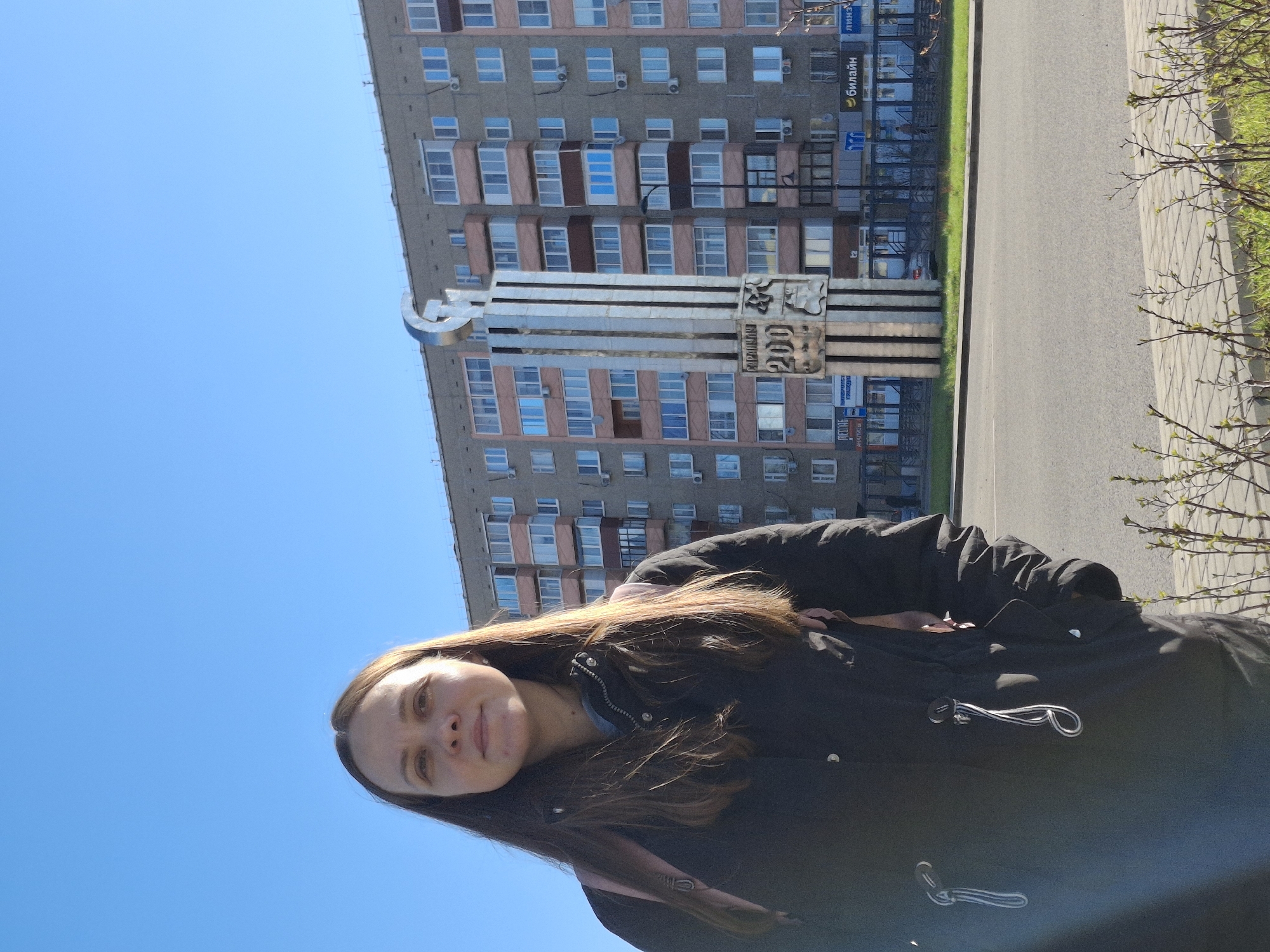 Here is a description of the image:
The image shows a young woman standing in front of a monument and a tall apartment building. The woman is wearing a dark winter coat and has long brown hair. The monument appears to be made of metal and features a "hammer and sickle" symbol. The apartment building behind the monument is tall and has many windows. The sky is blue and clear. The location is Unknown, Unknown.