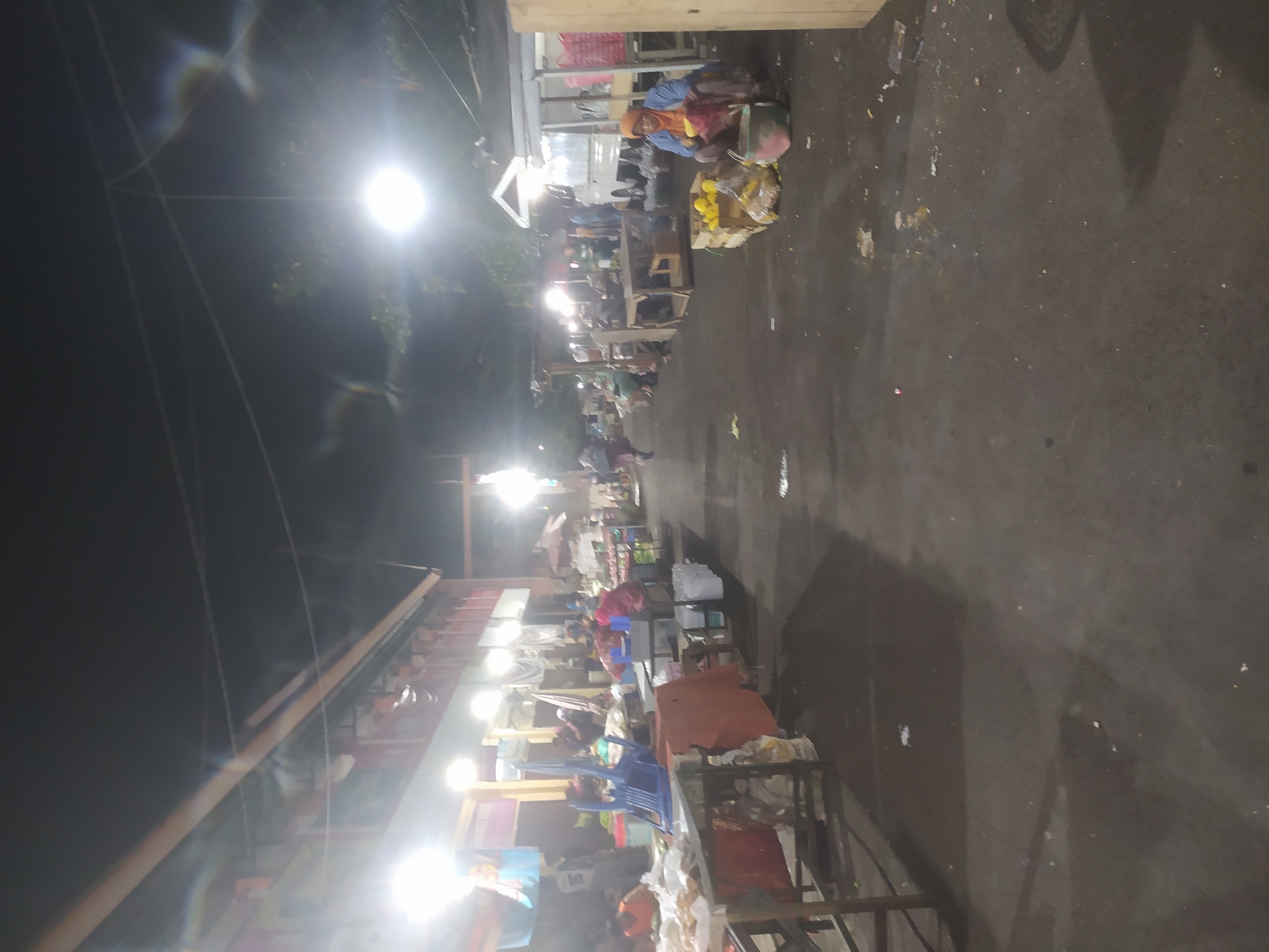 The image depicts a nighttime scene at a market in Kupang, Indonesia. The market appears to be an outdoor or semi-outdoor establishment, with stalls and vendors set up along a paved pathway. Bright lights illuminate the area, casting shadows and creating a slightly hazy atmosphere.
On the left side of the image, there are several stalls with tables and what appear to be goods displayed. Some people are visible behind the tables, possibly vendors. A blue plastic chair is prominently placed on one of the tables. Further down the row of stalls, more people are seen, some interacting with vendors.
In the center of the image, the pathway is visible, with some debris scattered on the ground. A few individuals are walking or standing in this area.
On the right side of the image, another vendor is seated next to a display of yellow produce, possibly corn, arranged in wooden crates. This vendor, wearing a blue top and orange headscarf, is engaged in an activity, though it is not clearly discernible from the image. A white truck is parked behind some of the stalls.
The overall environment suggests a bustling marketplace operating during the evening hours. The buildings in the background appear to be market structures or shops. The lighting suggests the presence of artificial light sources such as lamps or string lights.