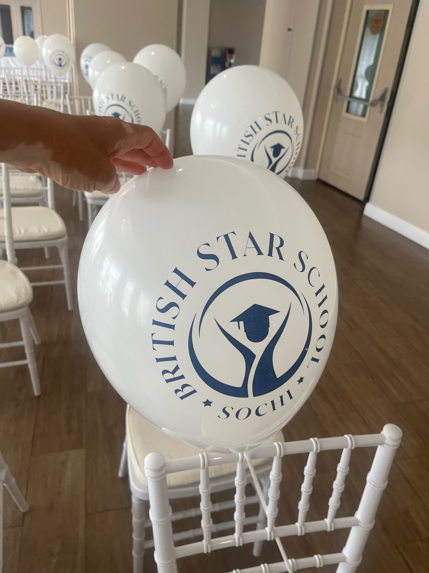 A person's hand is visible in the foreground, with vitiligo on the skin. The hand is reaching towards a large, white balloon. The balloon has a blue circular logo with text that reads "BRITISH STAR SCHOOL" and "SOCHI." The background shows a room with rows of white chairs, suggesting an event or gathering. In the far background, a door with a glass panel is visible, and part of a wall with a picture frame. The flooring is wooden. The image does not provide specific details about the exact location within Sochi, Russia, beyond the text on the balloon.