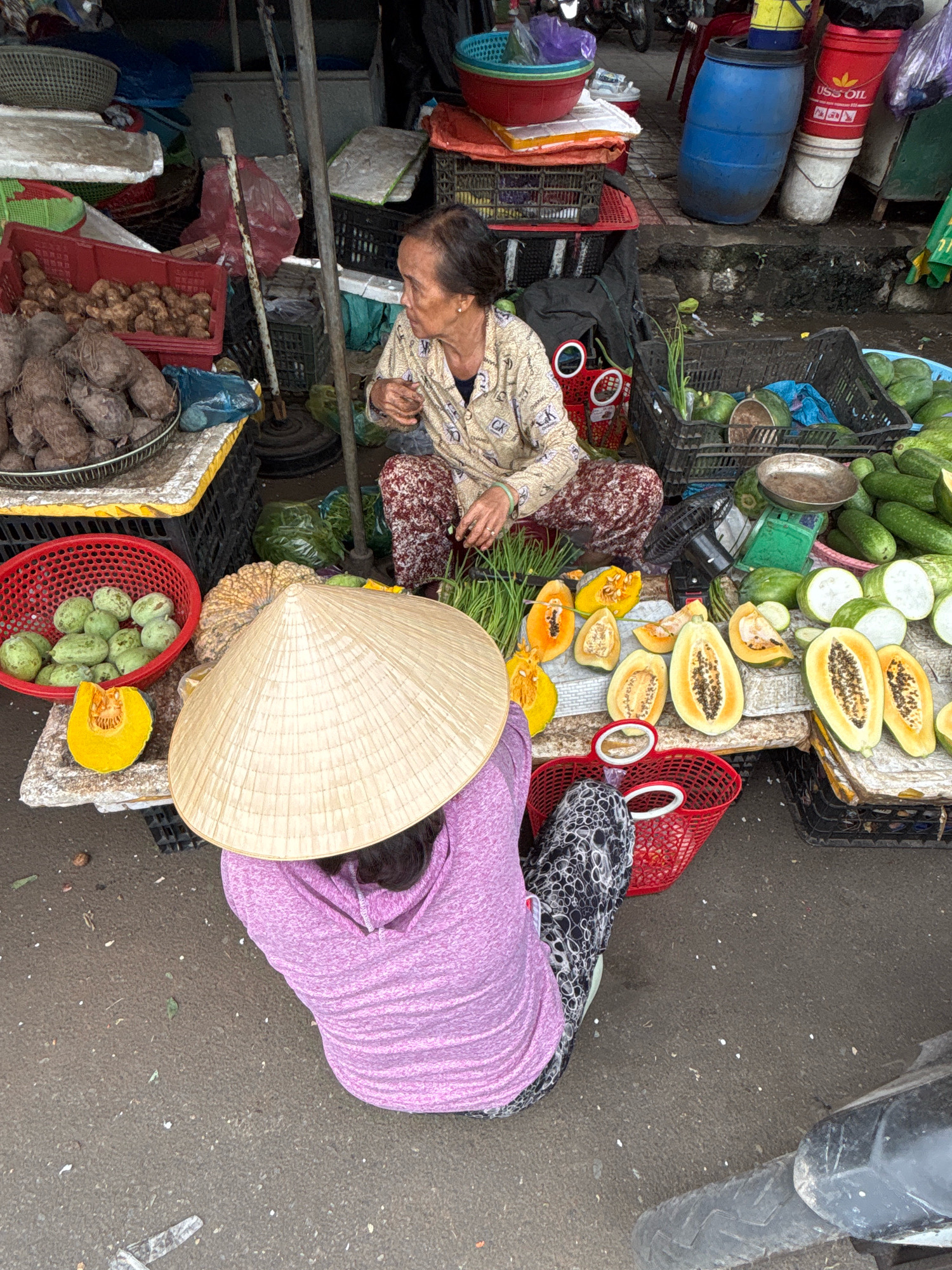 In Nha Trang, Vietnam, a busy street market scene unfolds. A woman, dressed in a patterned shirt and pants, sits on the ground tending to a display of fresh produce. In front of her, a variety of fruits and vegetables are neatly arranged on a table. Among them are sliced papayas, green gourds, and a whole pumpkin. A large conical hat, a traditional Vietnamese headwear, rests on the head of another individual, whose back is to the camera, and who is also seated on the ground. The market stalls are laden with goods, including baskets of roots, more produce, and various containers. The ground beneath is paved, and a portion of a parked motorcycle is visible in the lower right corner, suggesting a lively commercial area in the city.