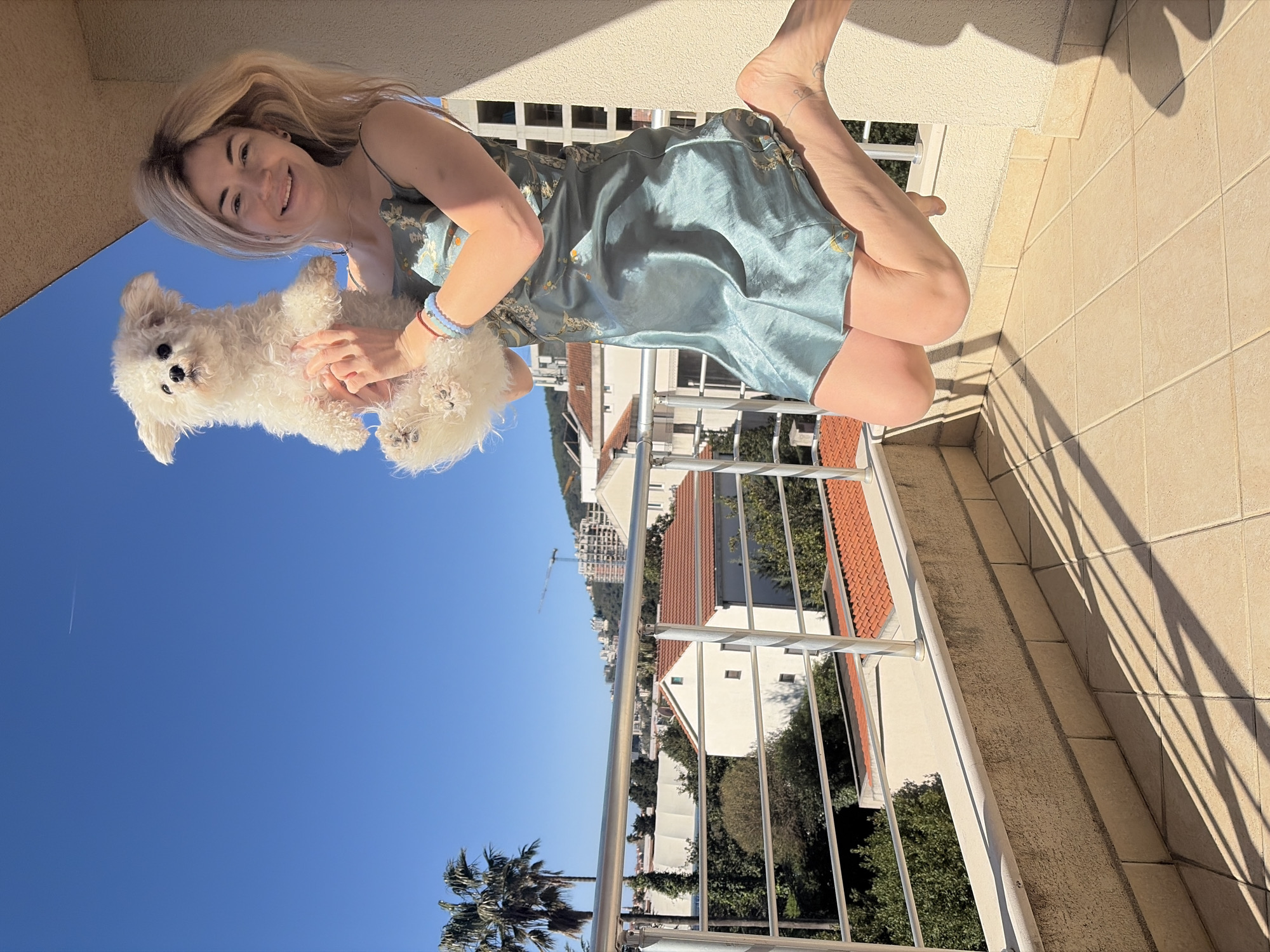 A young woman is holding a white, fluffy dog on a balcony in Budva, Montenegro, on a sunny day. She is wearing a short, green, patterned dress and is barefoot. She is looking at the camera and smiling. The dog is looking slightly to the left. Behind the balcony railing, there are houses with orange tile roofs, trees, and a view of the city. A palm tree is visible in the lower left corner of the frame. The sky is blue.