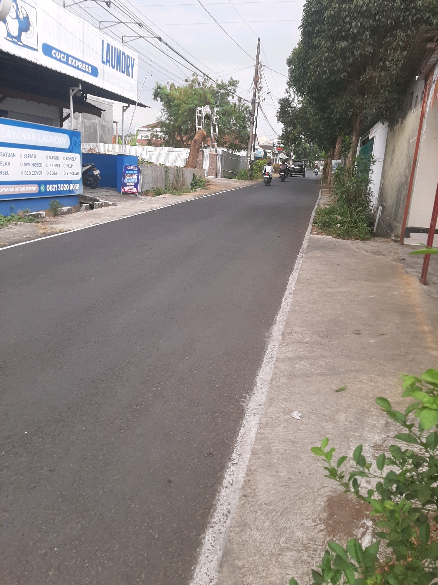 A street scene in Sumampir, Indonesia, features a road with a white line marking its edge. To the left of the road, a building with a "LAUNDRY CUCI EXPRESS" sign is visible, displaying various laundry services. A motorcycle is parked near the building. On the right side of the road, a concrete sidewalk runs alongside a wall and trees. A few motorcycles and a car are on the road, with people riding them, suggesting ongoing activity. The sky is overcast.