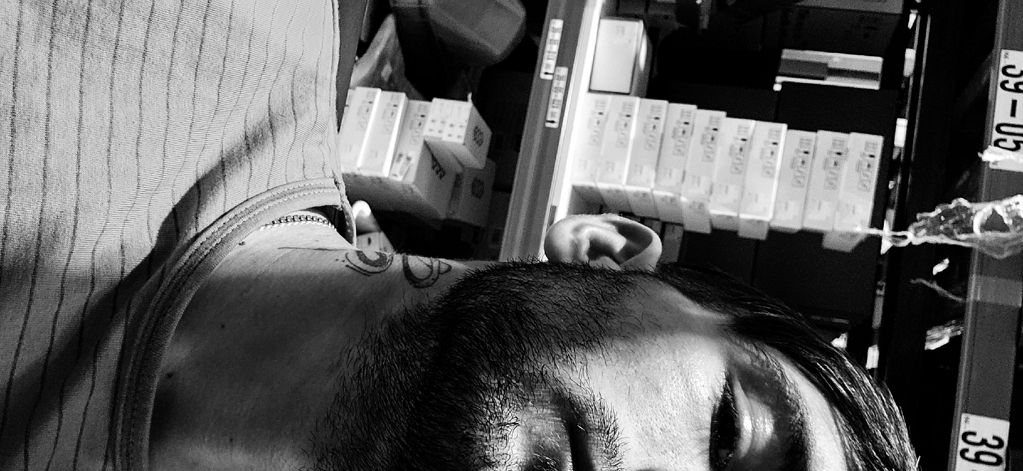 This black and white image is a rotated portrait (90 degrees clockwise) featuring a person, likely taken in Plovdiv, Bulgaria. The subject's head, face (partially obscured), shoulder, and upper arm are visible in the foreground. They are wearing a ribbed tank top or shirt with thin, vertical stripes. A necklace with small, interlocking links or beads is visible around their neck. On their left shoulder or collarbone area, there is a distinct tattoo composed of two stylized, possibly eye-like, shapes.
The person's dark hair is visible, and their eyes appear closed or looking downwards, suggesting a moment of rest or contemplation. Stubble is visible on their chin and jawline.
In the background, which is out of focus, rows of white boxes are stacked on shelves. This suggests an indoor setting such as a storage room, warehouse, or the backroom of a retail establishment. A white label with the text "39-05" and "39" is visible on a vertical surface on the right side of the frame, possibly attached to a shelf or a column. The overall lighting appears soft, creating subtle shadows on the person's clothing and face.