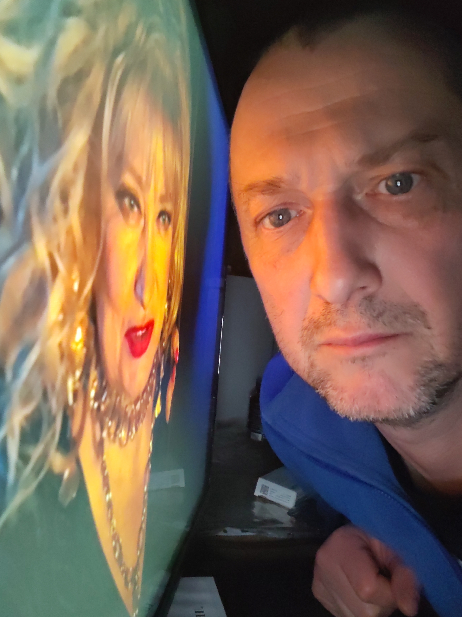 The image, taken in Shcheleiki, Russia, presents a close-up view of a man looking towards a screen displaying a woman. The man, positioned on the right side of the frame, has short hair, light stubble, and visible lines on his forehead. He wears a blue jacket or hoodie and gazes towards the screen with a neutral expression.
On the left, a bright screen, likely a television or monitor, shows a woman with voluminous blonde, curly hair, pronounced red lipstick, and prominent jewelry, including a large necklace and earrings. She appears to be wearing a green outfit. The screen emits a strong, warm light that illuminates both the woman's face and casts an orange glow on the man's right cheek and side of his face.
The setting appears to be indoors and dark, suggesting evening or night, with the primary light source being the screen. In the background, on a dark surface beneath the screen, a small white box with a QR code is visible. Below it, a piece of paper shows partial text, possibly "IL". The composition highlights the man's direct engagement with the content on the display.