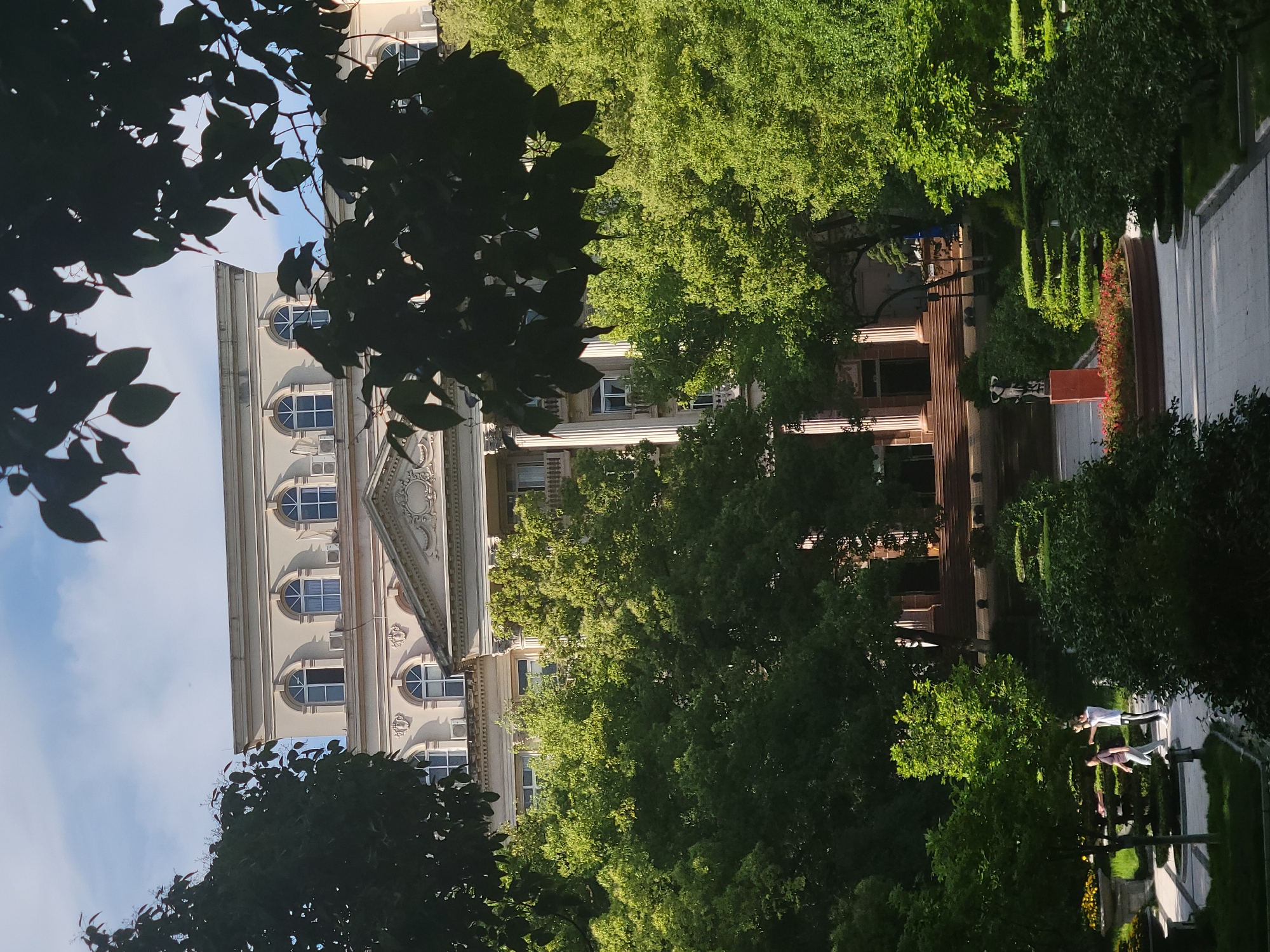 A low-angle, partially obscured view reveals a building with arched windows and decorative elements. Foliage frames the view. Further back, a walkway is seen with several people on it. The location is Beibei, China.