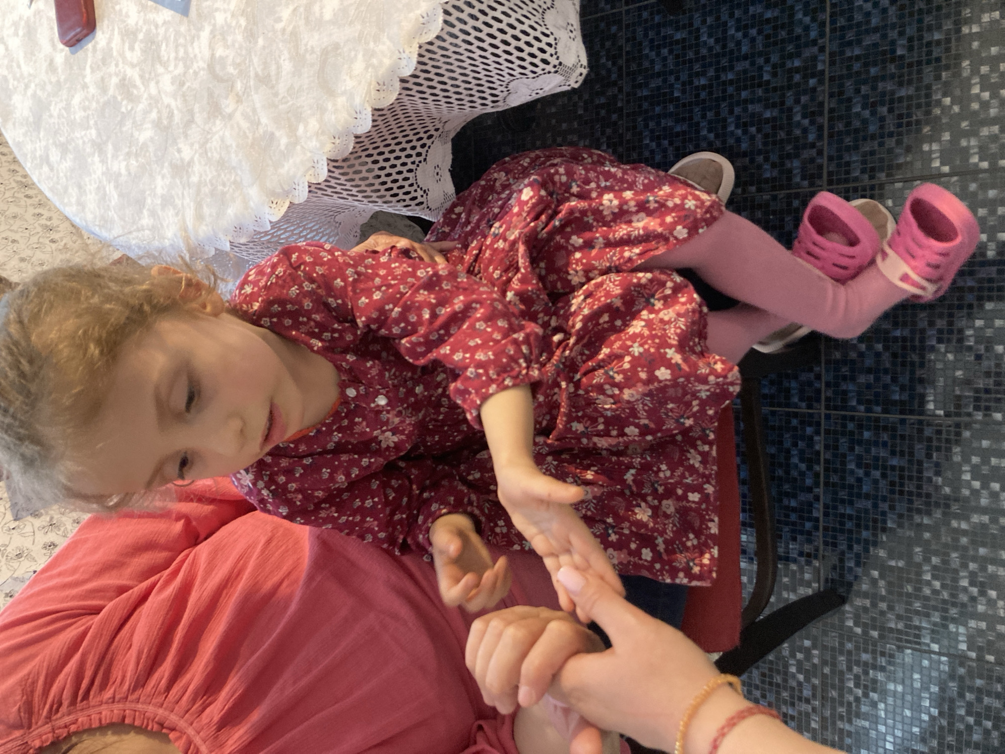 A high-angle shot depicts a young girl with curly blonde hair sitting on a woman's lap. The girl is wearing a red floral dress and light pink leggings. On her feet, she wears bright pink clogs on one foot and a tan colored shoe on the other. The woman is wearing a salmon colored top and has her right arm extended, clasping the girl's hand. The woman is wearing gold and red bead bracelets on her right wrist. A white lace tablecloth covers a nearby table. The floor is tiled with small blue and black squares. The location is Unknown, Unknown.