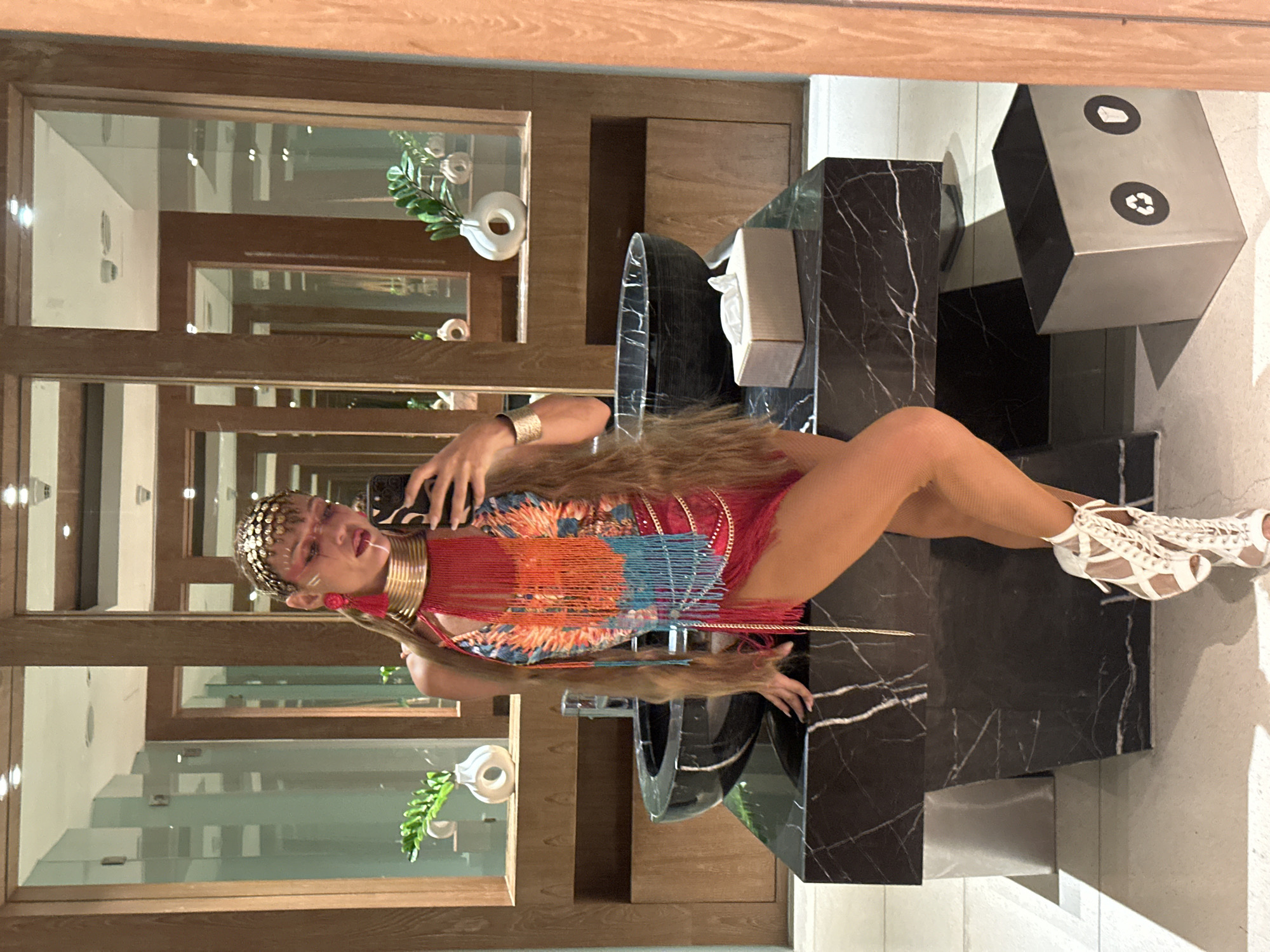 A woman in a vibrant, fringed outfit and elaborate headpiece stands in a modern bathroom, taking a selfie. The bathroom features black marble countertops, a large black sink, and mirrors reflecting the space. A tissue box sits on the counter, and a metal trash can with recycling symbols is visible on the floor. The woman's hair is long and blonde, and she is wearing white, strappy heeled sandals. The background suggests an upscale establishment, possibly a hotel or resort in a tropical location like Ko Samui, Thailand, given the plants and overall aesthetic.