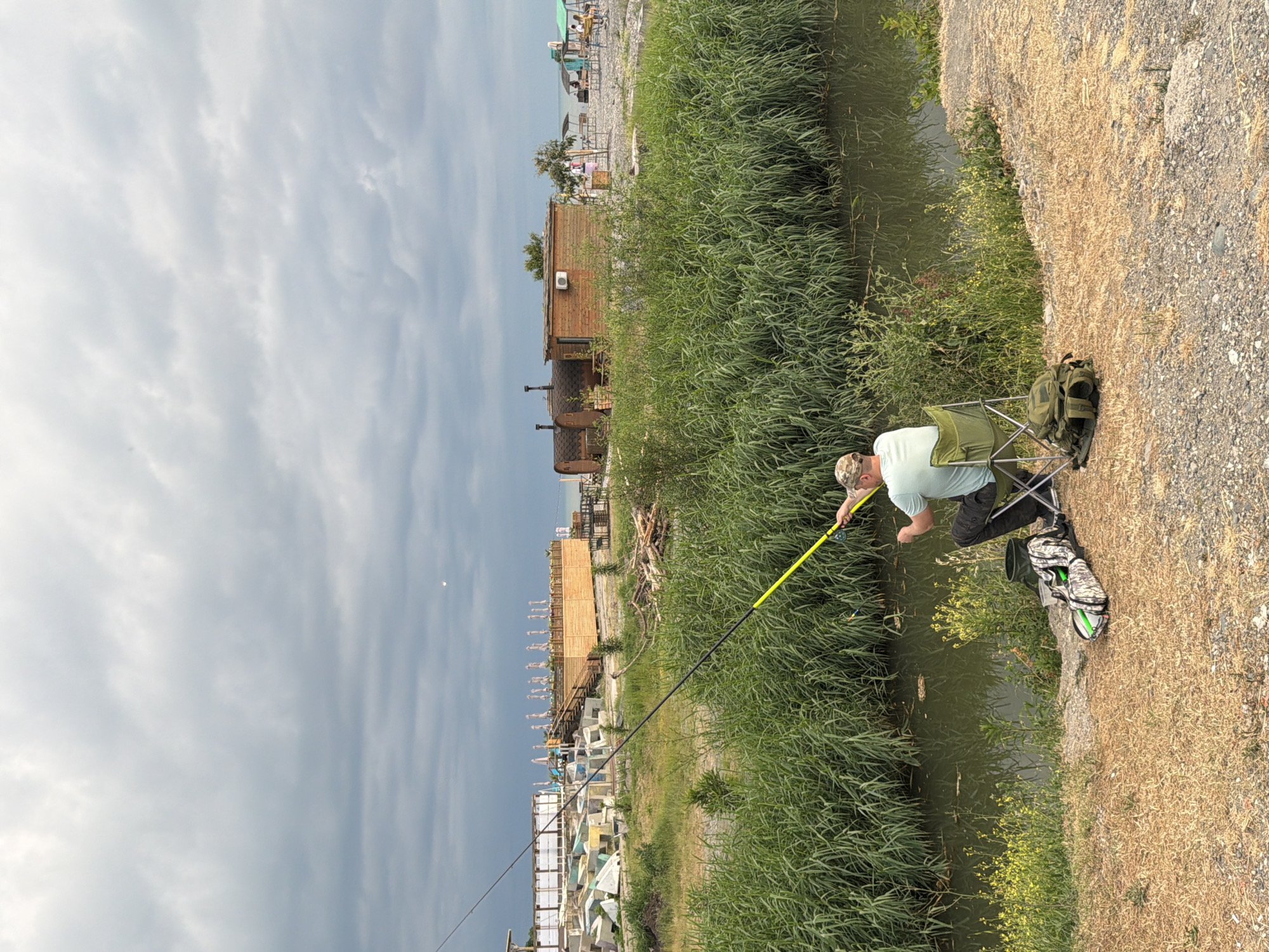 The image depicts a man fishing on the bank of a narrow canal. He is seated in a folding chair, holding a fishing rod with a yellow tip. A camouflage backpack rests on the ground next to him. The man is wearing a light-colored t-shirt and a camouflage cap. The canal is bordered by tall, green reeds on one side and a dry, gravelly bank on the other. In the background, there are several wooden structures, possibly saunas or small buildings, and a beach area with umbrellas and lounge chairs, suggesting a resort or recreational area. The sky is overcast with dramatic, heavy clouds, indicating a potentially stormy or moody weather. The overall scene appears to be set in Sochi, Russia, given the provided location context. The lighting suggests it might be late afternoon or early evening. The man's posture and focus on fishing imply a moment of calm and concentration amidst the natural and man-made elements of the scene.