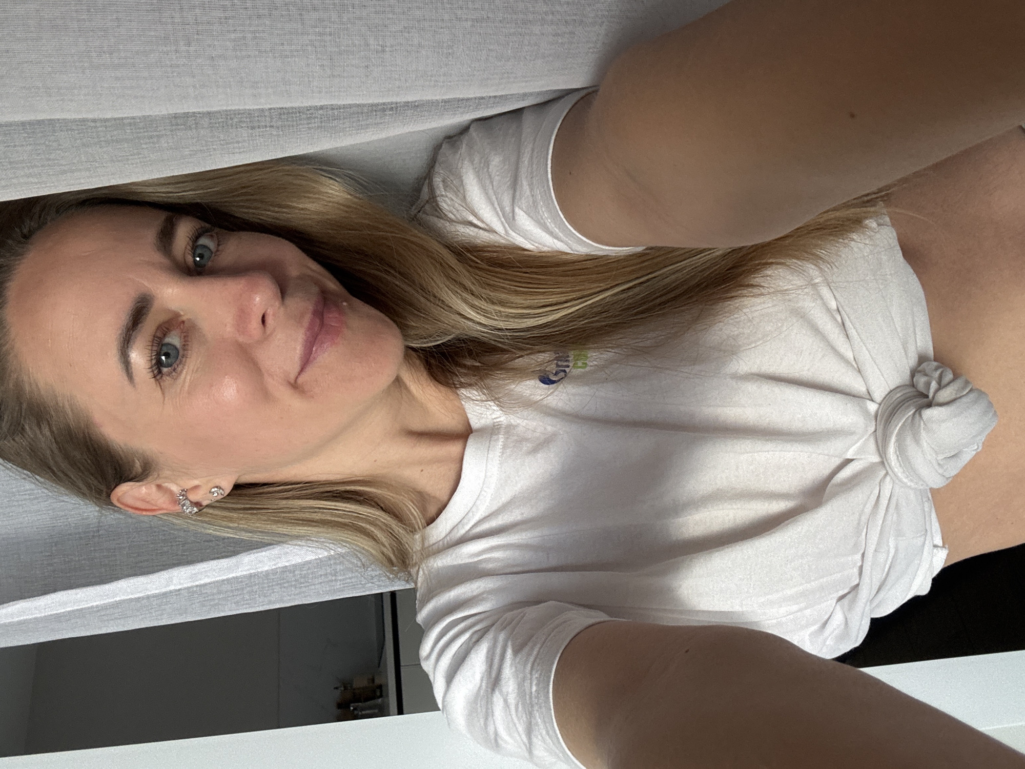 A woman with blonde hair and blue eyes is taking a selfie. She is wearing a white t-shirt tied at the waist and a silver earring. She is standing near a white curtain. The background shows a kitchen area. There is no information that can determine the location in the city of Chaikovsky, Russia.