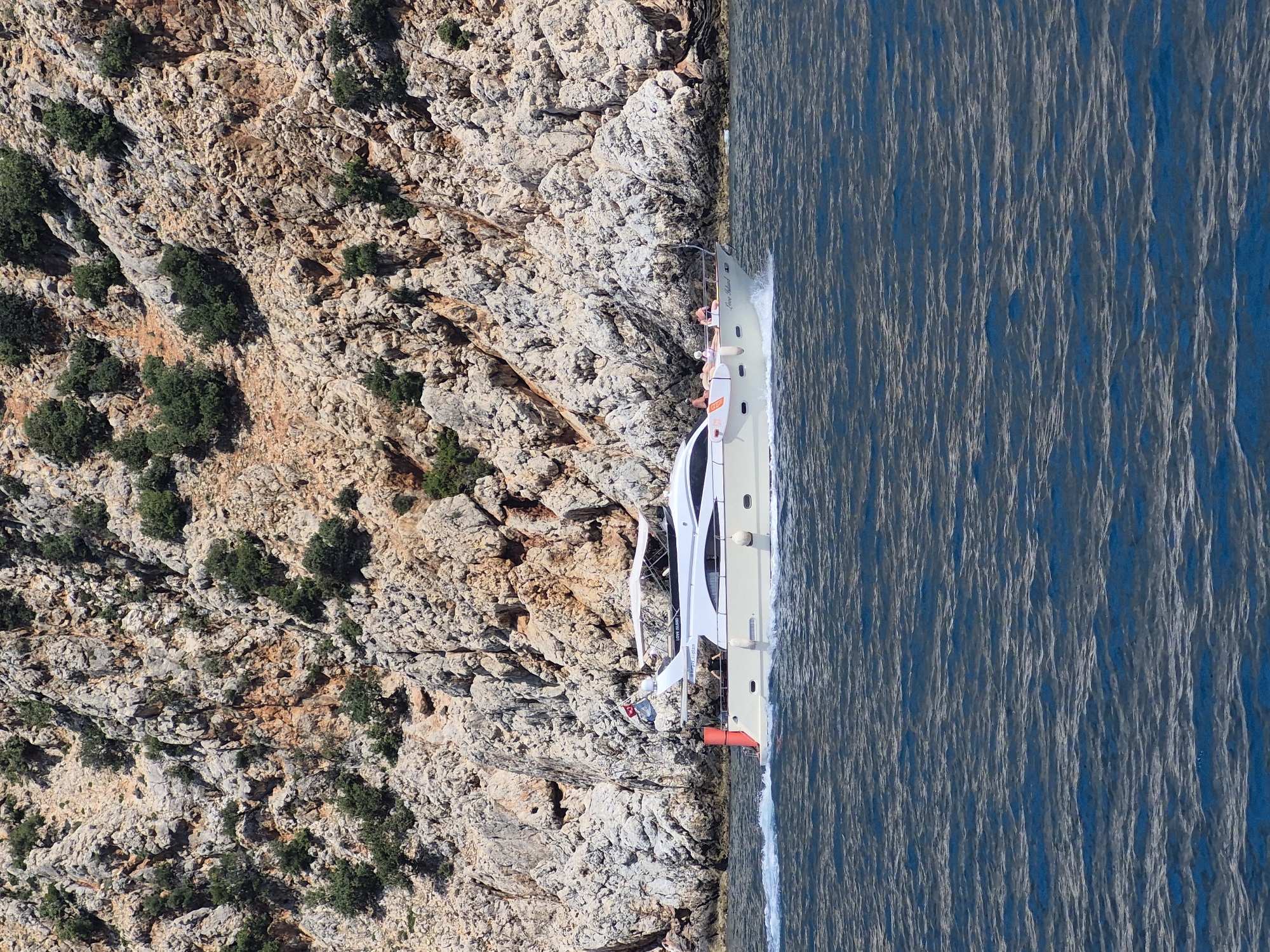 Aerial view of a boat near the rocky coast of Alanya, Turkey. The boat is cruising on the water, close to the edge of a rugged rock formation. The rock is a mix of gray and brown hues, dotted with patches of green vegetation. The water is a deep blue, textured with wave patterns. The boat, carrying several people, is light-colored and appears to be moving at a moderate speed, evidenced by the white wake trailing behind it. The angle of the shot suggests it was taken from above, possibly from a drone or a high vantage point.