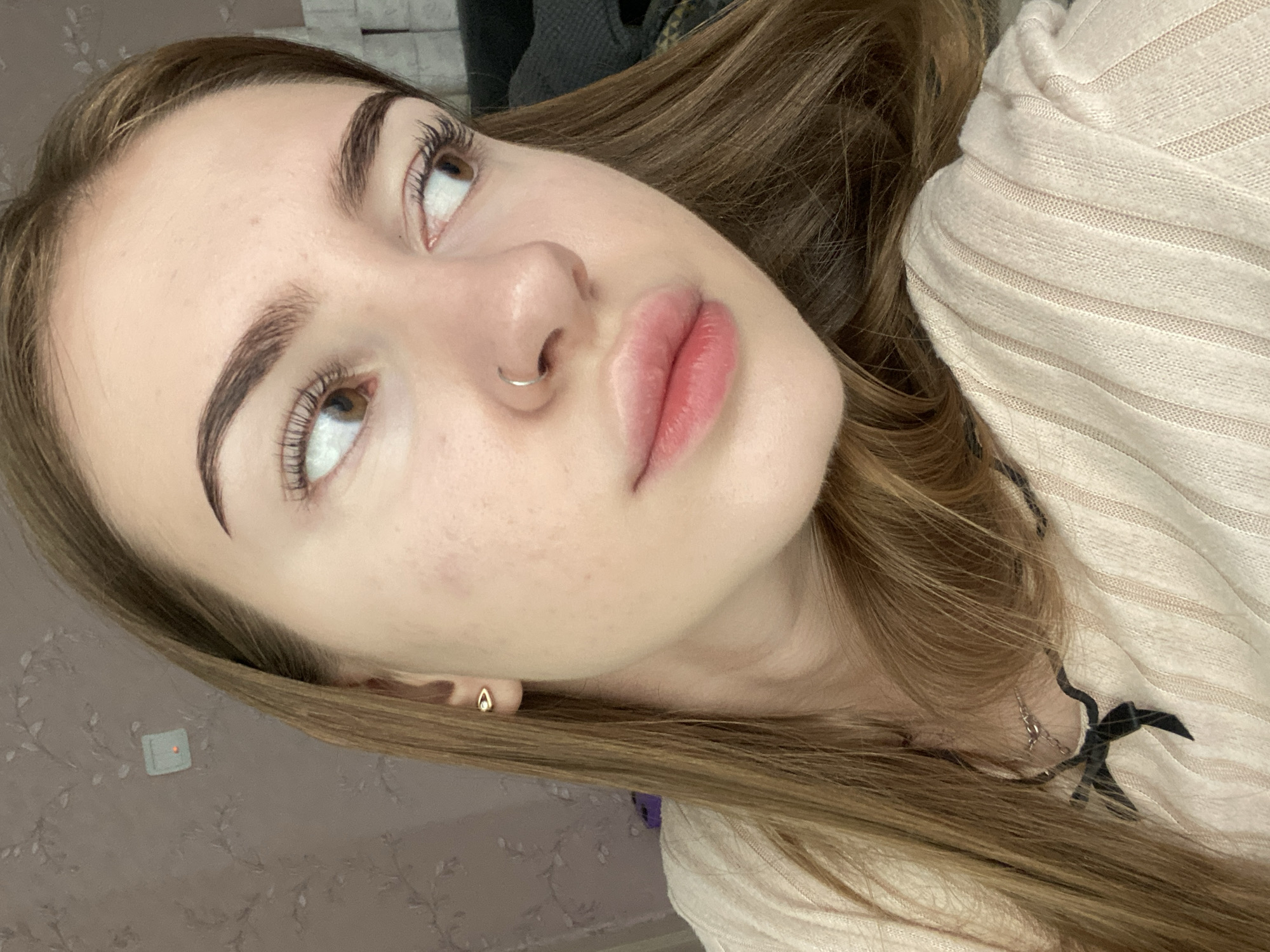 A close-up, head-and-shoulders selfie, taken indoors against a light-colored, floral-patterned wallpaper, shows a young Caucasian woman with long, light brown hair. She is looking to her right, her gaze averted from the camera. Her eyebrows are well-defined and dark. She has large, brown eyes, accentuated by mascara. A small, gold, tear-drop shaped earring adorns her left earlobe, and a silver hoop pierces her right nostril. Her lips are full and slightly parted, appearing to have a subtle pink tint. She is wearing a cream-colored, ribbed knit sweater with thin vertical stripes. A delicate silver necklace with a dark bow charm hangs around her neck. The lighting is soft, suggesting an indoor environment, possibly a home. There is no information to determine the location as Дьяково, Russia.