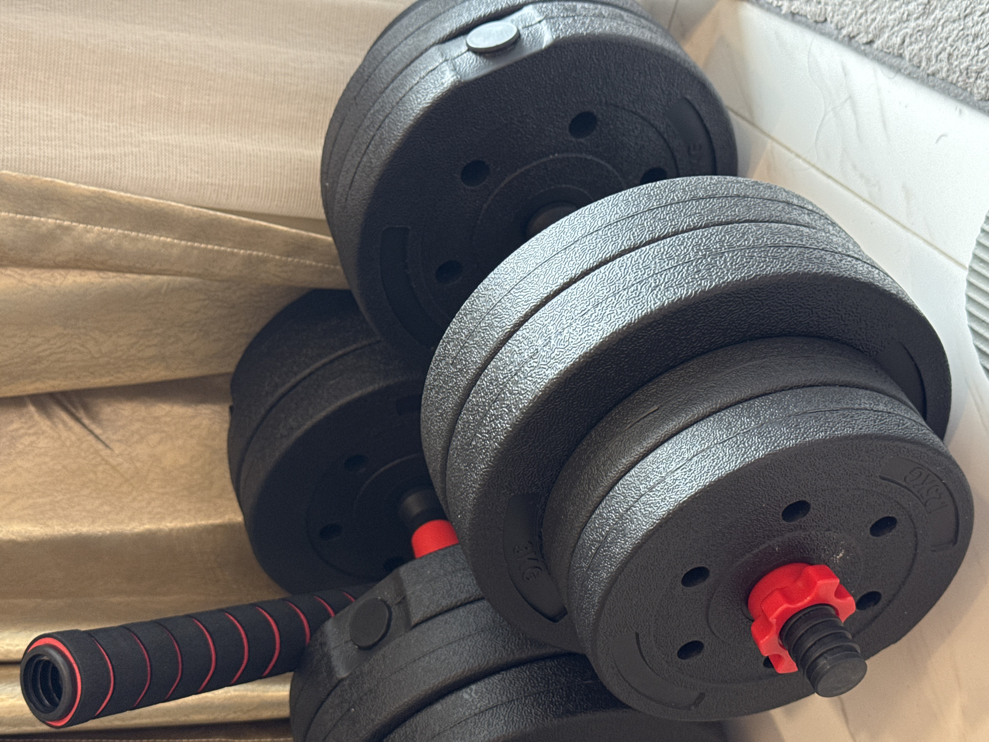 This image features two adjustable dumbbells and their components, set in what appears to be an indoor environment. The primary subjects are the exercise weights, consisting of multiple black, textured weight plates and two dumbbell handles.
One assembled dumbbell is prominently displayed in the foreground on the right, with several black plates secured by a bright red, star-shaped screw-on collar. Another set of weight plates is visible in the background, partially assembled on a handle. The plates have a matte, slightly bumpy finish and feature small circular holes. Visible text on some plates clearly indicates their weight, specifically "2.5KG" and "1.25KG."
One black dumbbell handle is positioned in the lower-left corner, featuring a black foam or rubber grip with distinct red rings providing ergonomic segmentation. The handle is unattached to the weights in this view but shows its design for comfortable handling. Parts of another handle, also with red accents, are visible where the weight plates are stacked.
The setting appears to be an interior space, with the dumbbells resting on two distinct surfaces. On the left and in the upper background, there is a light beige or cream-colored upholstered fabric, possibly a couch or bed. On the right, the weights are partially on a white tiled floor, next to which a grey textured rug or mat is visible. The lighting suggests an indoor daytime or well-lit evening setting. There are no people visible, and no activity is taking place beyond the static arrangement of the gym equipment. The image provides no specific visual cues to indicate the location context of Pattaya City, Thailand.