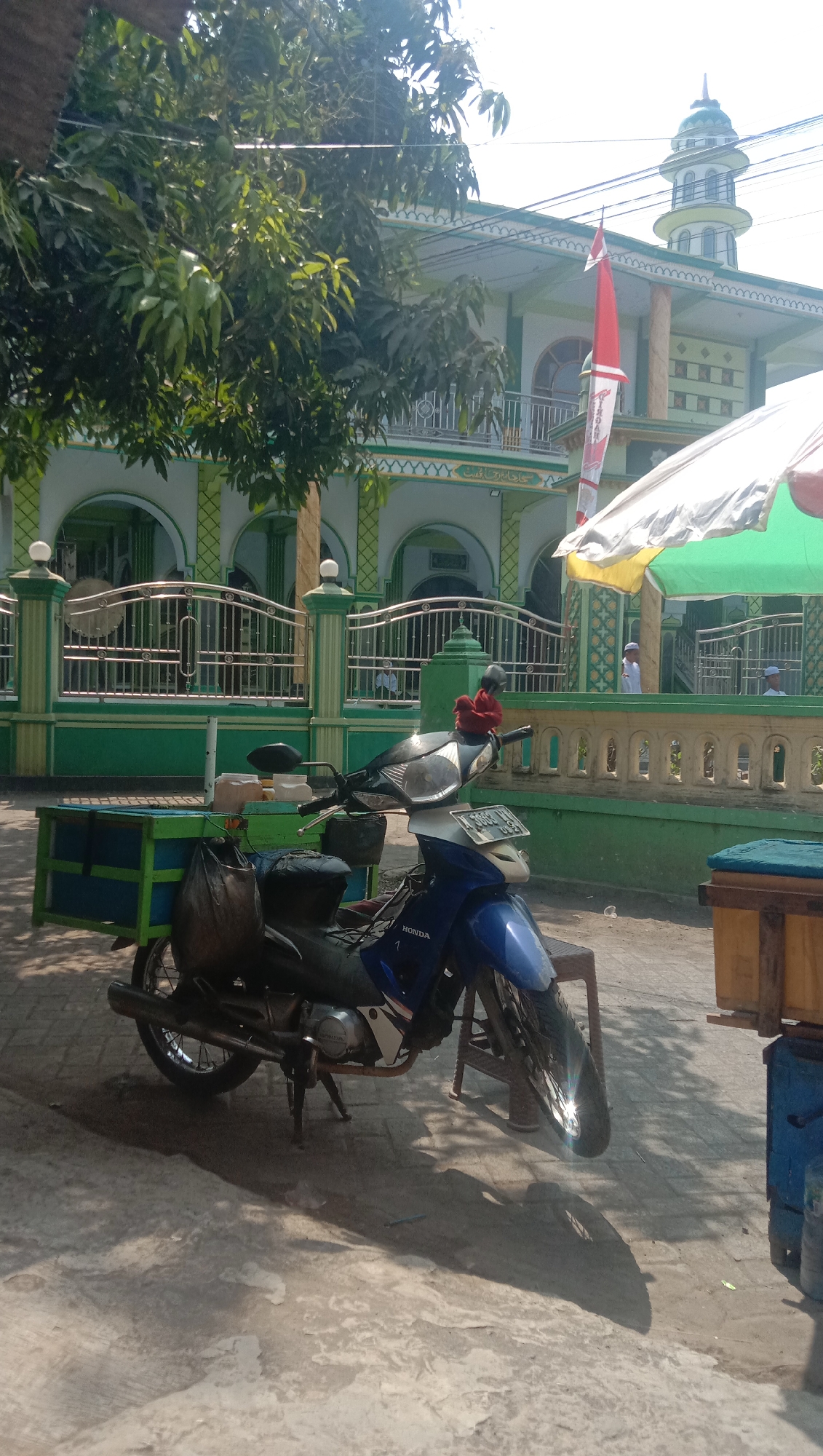 In the city of Ngempit, Indonesia, a blue and silver Honda motorcycle is parked on a concrete surface in front of a mosque. The mosque has a green and white facade with arched windows and a minaret topped with a green dome. A red and white flag is displayed near the mosque's entrance.
The motorcycle is equipped with a green and blue wheeled cart attached to its rear. A black bag hangs from the cart. The motorcycle's handlebars are adorned with a red fabric decoration. A white and yellow umbrella is positioned to the right of the motorcycle, providing shade. In the background, several people, possibly mosque attendees, are visible wearing white attire. The scene is bathed in sunlight, casting shadows on the ground. A tree with dense foliage stands to the left of the mosque, partially obscuring the view of the building. The overall environment suggests a public outdoor space.