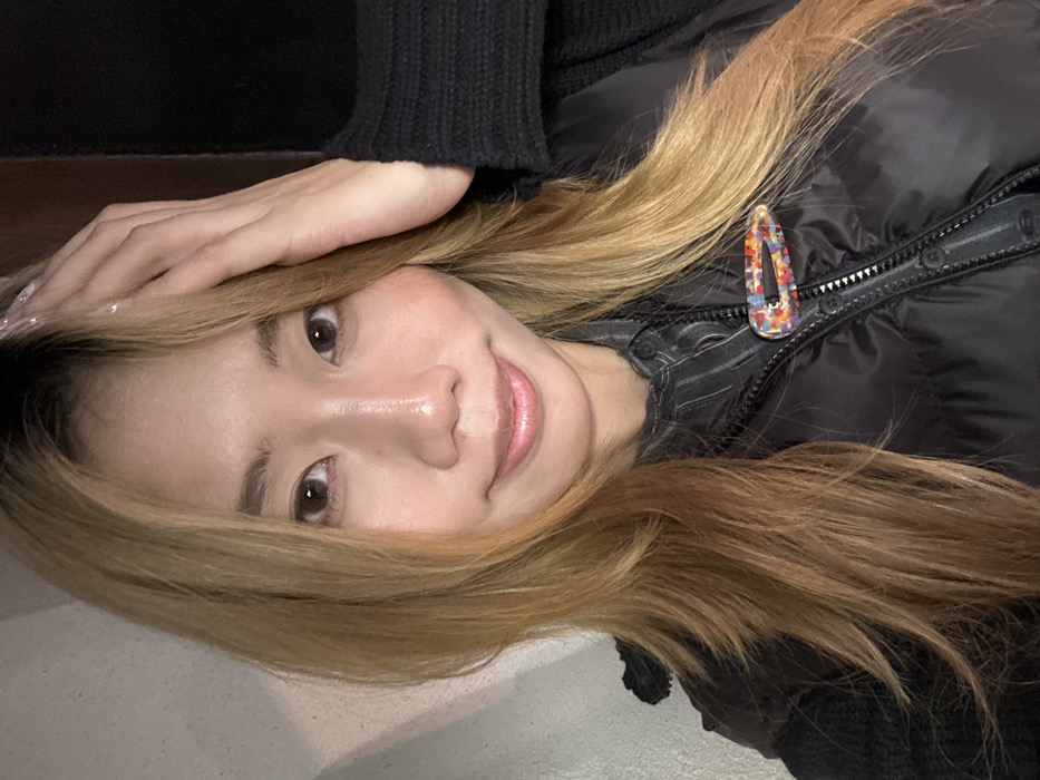 An eye-level portrait depicts an East Asian female in the city of Liangjiang, China. She possesses light brown, layered hair with visible dark roots, extending to her shoulders. Her face is oriented forward, meeting the camera's gaze with a subtle smile. Her left hand, displaying clear-coated fingernails, rests on the crown of her head, fingers slightly spread. She wears a black puffer vest, partially zipped, over a dark, subtly striped collared shirt. A black knit sleeve is visible on her right side. A colorful, speckled barrette, exhibiting red, orange, yellow, and blue hues, is clipped to the left side of her vest. The background is a uniform, light-toned wall surface.