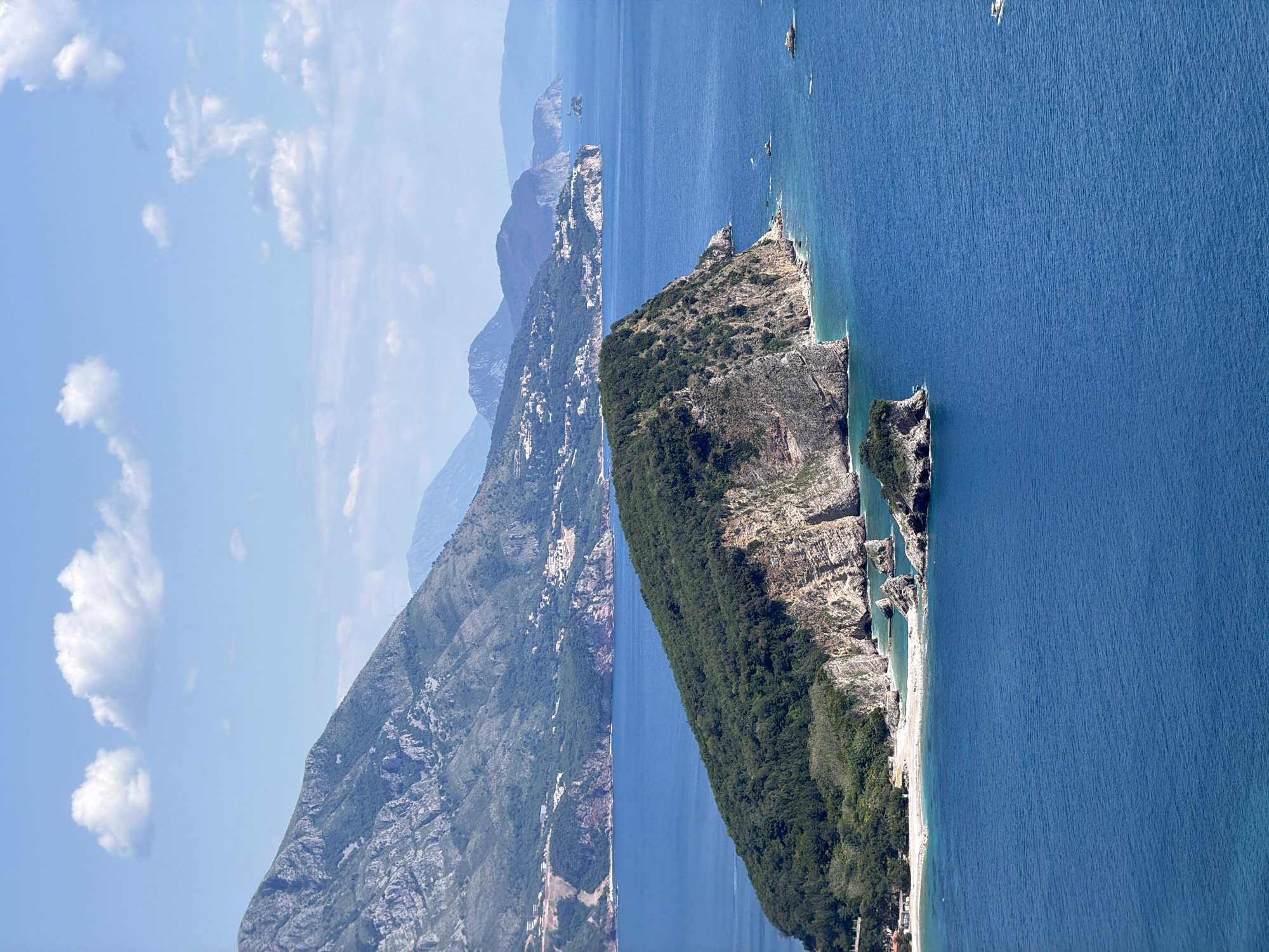 The image shows a high-angle view of the coastline near Budva, Montenegro. In the foreground, a rocky, tree-covered peninsula extends into the blue sea. A small beach is visible along the peninsula's edge. Across the water, a mountain range rises sharply, its slopes dotted with buildings and vegetation. The sky is blue with scattered white clouds. Several boats are visible on the water.