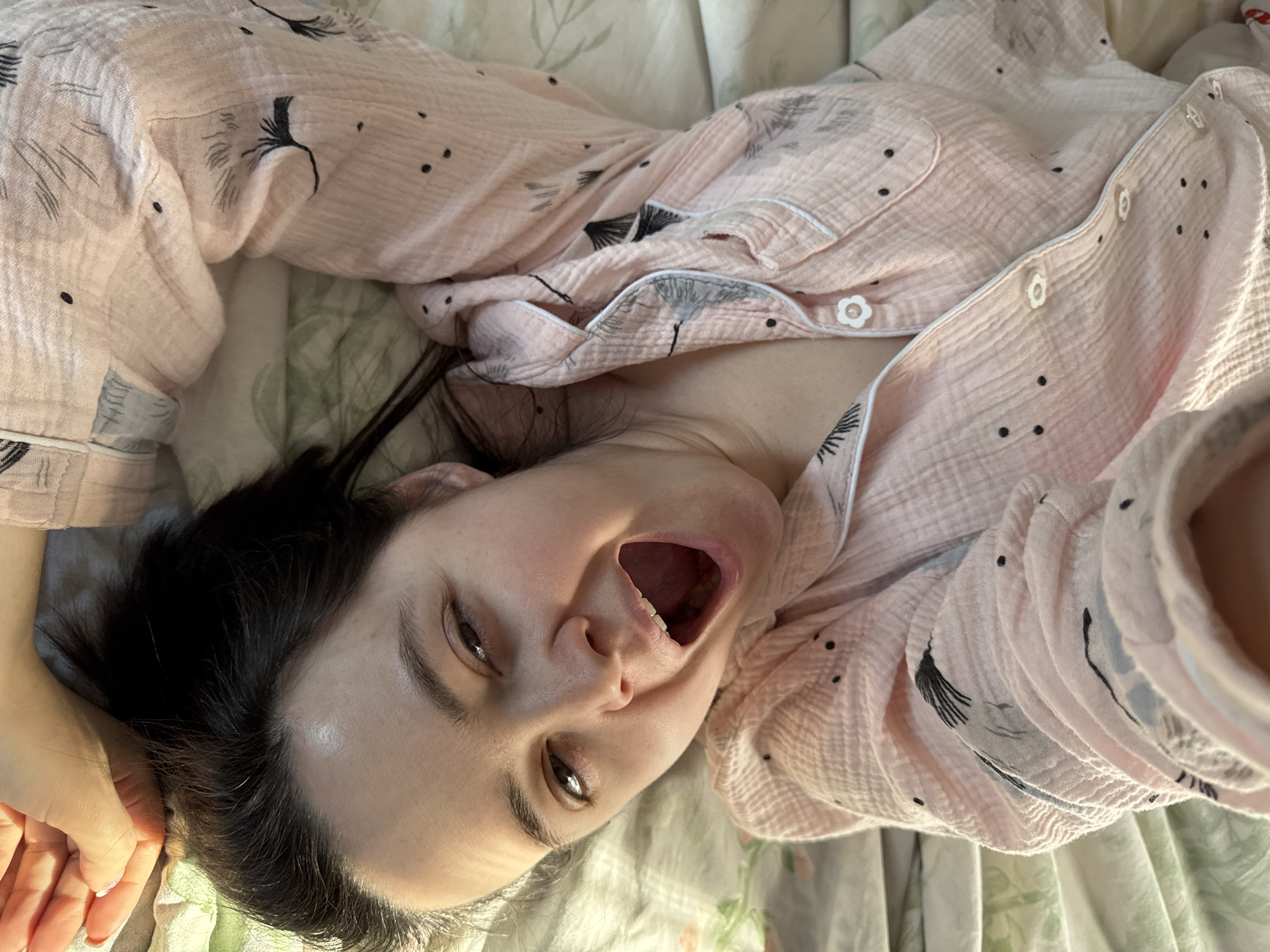 A woman lies supine on a bed with her face turned towards the camera. She is wearing a pink button-down pajama top with a pattern of small black dots and stylized leaf-like designs. Her mouth is open in what appears to be a yawn or shout, and her dark hair is disheveled around her head. The bedding beneath her has a floral pattern. Her left hand is visible near her head, and the overall lighting suggests a daytime setting. There is no information in the image to indicate it was taken in Izhevsk, Russia.