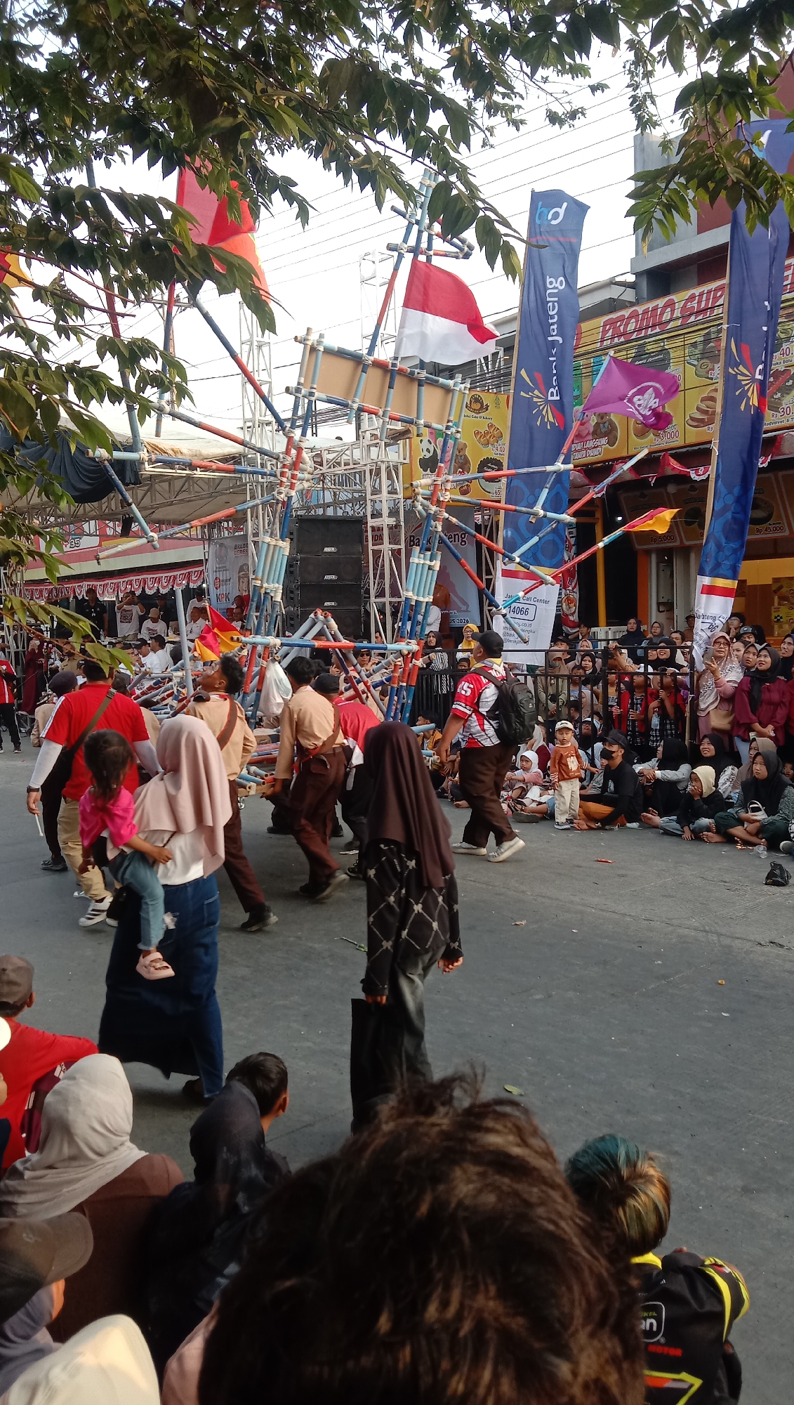 The image depicts an outdoor street scene in Jatisawit, Indonesia, characterized by a vibrant parade or festival atmosphere. In the foreground, a group of people are gathered, with some seated and others standing. The majority of the people are facing away from the camera, towards the action unfolding in the mid-ground.
The mid-ground is dominated by a colorful, multi-tiered structure constructed from bamboo and scaffolding, suggesting it's part of a performance or float. Several individuals, some dressed in what appear to be school uniforms (khaki shirts and brown pants), are actively engaged around this structure, carrying or maneuvering parts of it. A prominent red and white Indonesian flag is displayed on a pole atop this structure. Adjacent to it, other flags, including a purple scout flag and blue banners, are visible.
Behind the central structure, a stage area with speakers and equipment is set up, indicating a performance venue. The background reveals a street lined with buildings, some of which have banners advertising businesses, including "PROMO SUPER" and "Bank Jateng". A crowd of spectators is positioned along the roadside, observing the event.
The overall environment suggests a daytime event, with natural light illuminating the scene. The presence of trees with foliage in the upper left corner frames the view. The ground is paved, indicating an urban or semi-urban setting. The people in the image are diverse in age, with children and adults participating and observing the festivities. The attire of the participants, particularly the uniforms, suggests an organized event, possibly a school or community celebration.