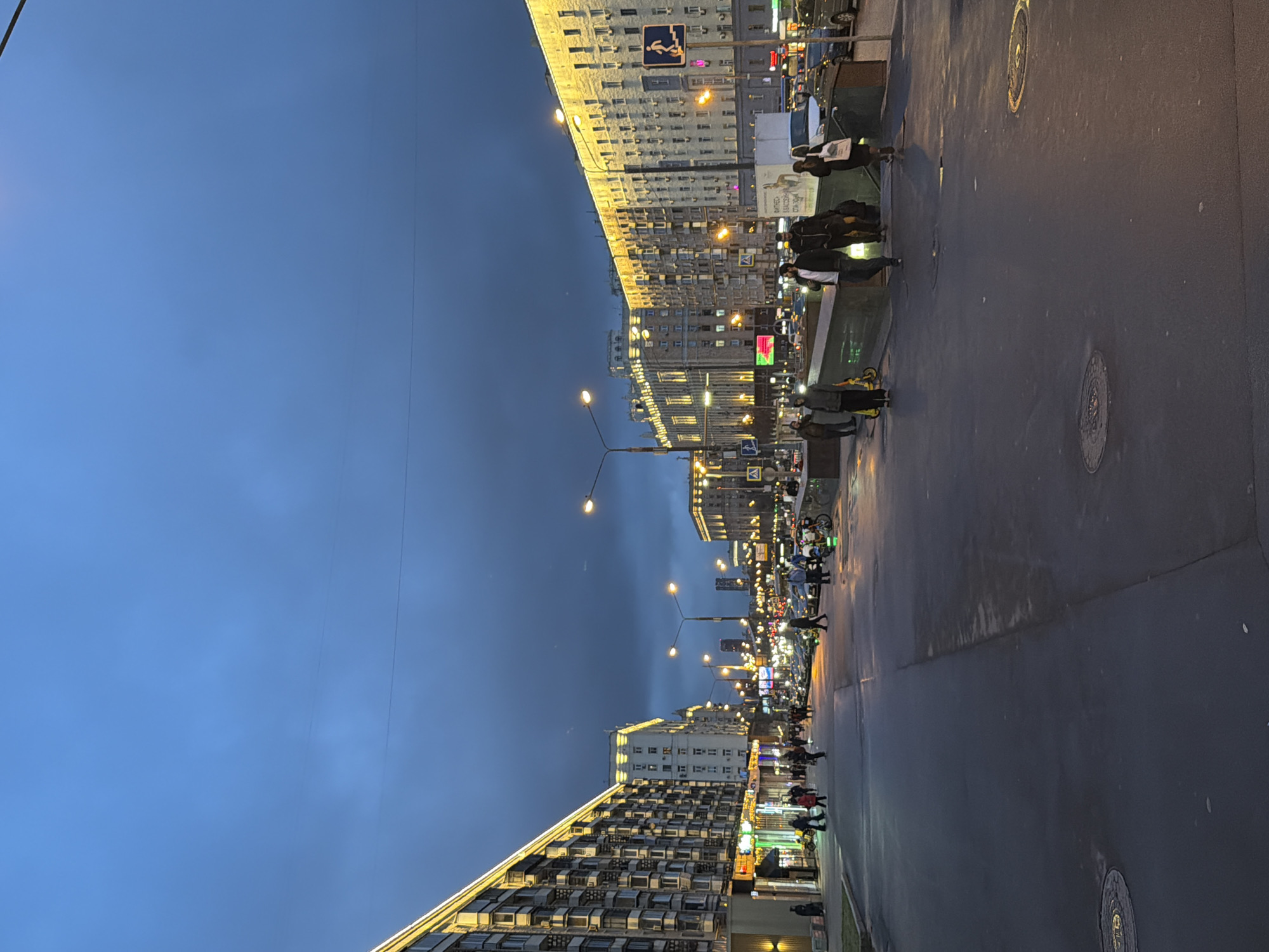 The image shows a street in Moscow, Russia, at dusk or twilight. Tall buildings line both sides of the wide street, illuminated with lights. Many people are walking along the sidewalks, and some are crossing the street. Streetlights illuminate the scene, and the sky is a deep blue color. The street surface appears wet, reflecting the lights.